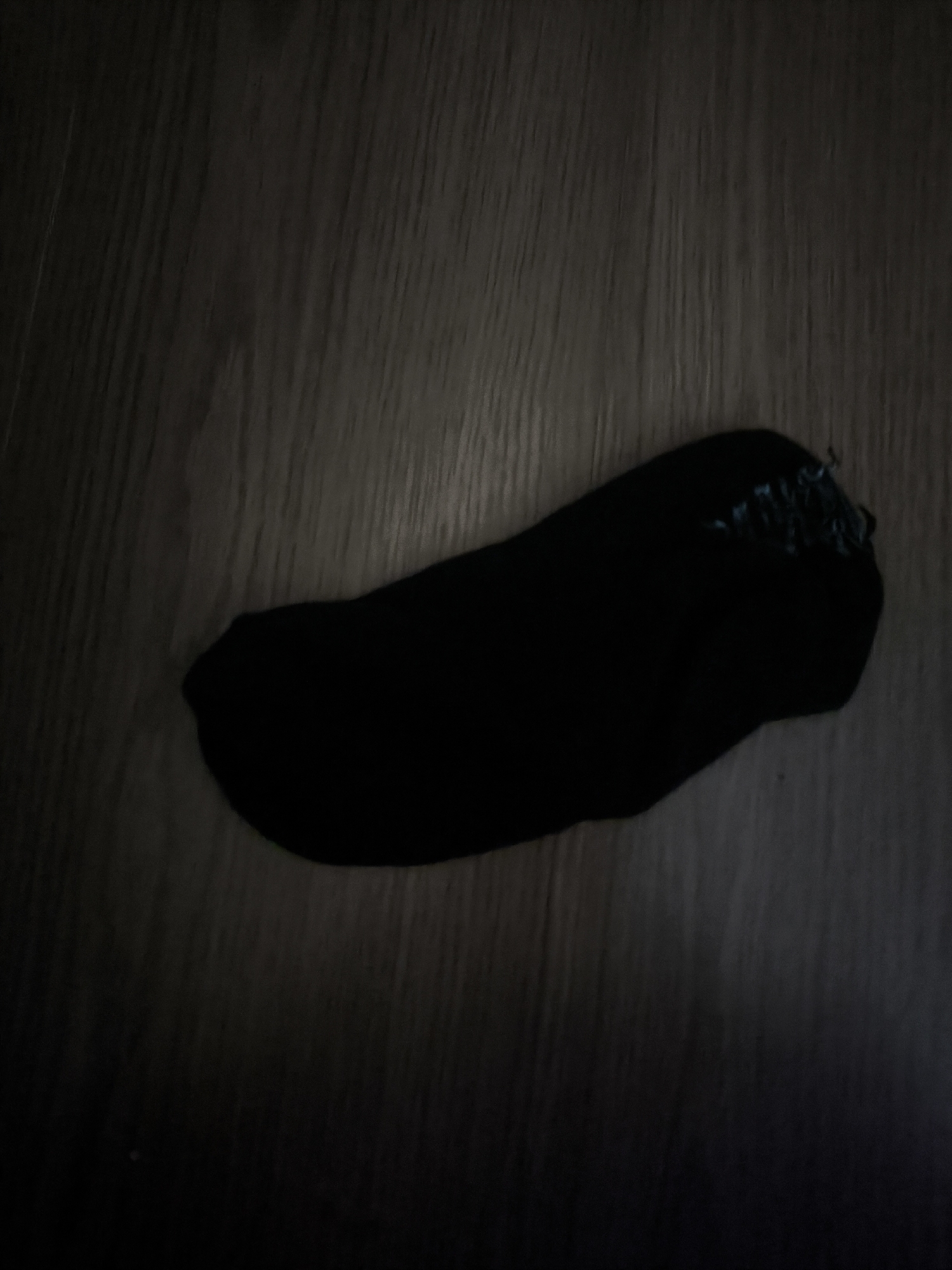 A black sock is lying on a dark wooden floor. The sock is positioned horizontally across the frame, with the toe pointing to the left and the heel to the right. The heel area appears slightly frayed, with some visible threads. The lighting in the image is dim, casting long shadows on the floor and obscuring some details. The background consists of wooden planks with a visible grain pattern, running vertically across the image. There are no people or other objects present in the scene. The setting is indoors, likely a residential area, but its specific location within Almaty, Kazakhstan, cannot be determined from the visual information provided.
