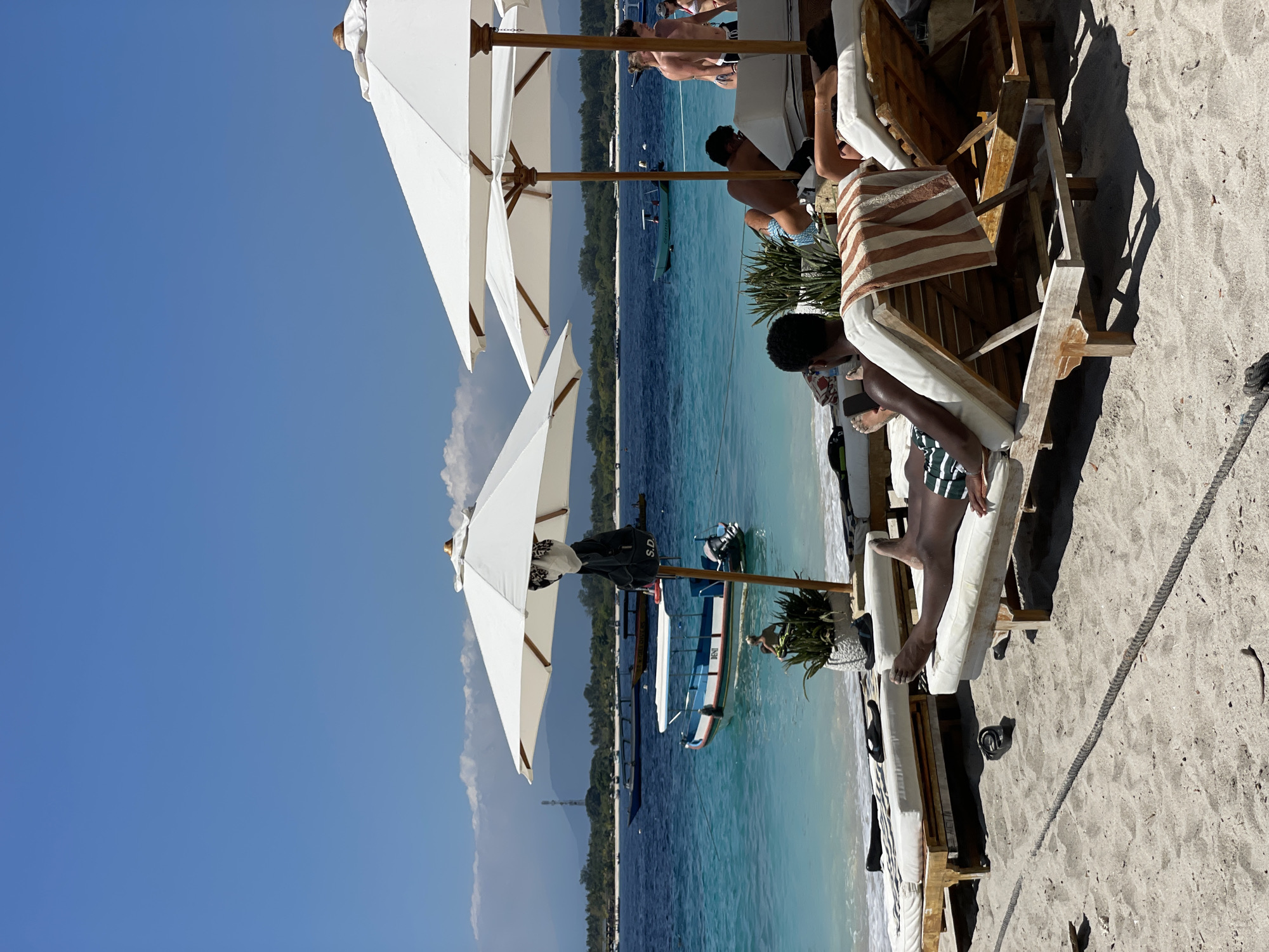 In Unknown, Indonesia, a serene beach scene unfolds under a clear blue sky. Two white umbrellas provide shade for lounge chairs on the sandy shore. In the foreground, several individuals recline on the chairs, some with towels draped over them. One person in the middle ground is partially visible, seated on a chair. Further in the background, the turquoise ocean stretches out, dotted with several boats. A distant shoreline with lush greenery and mountains forms the horizon.