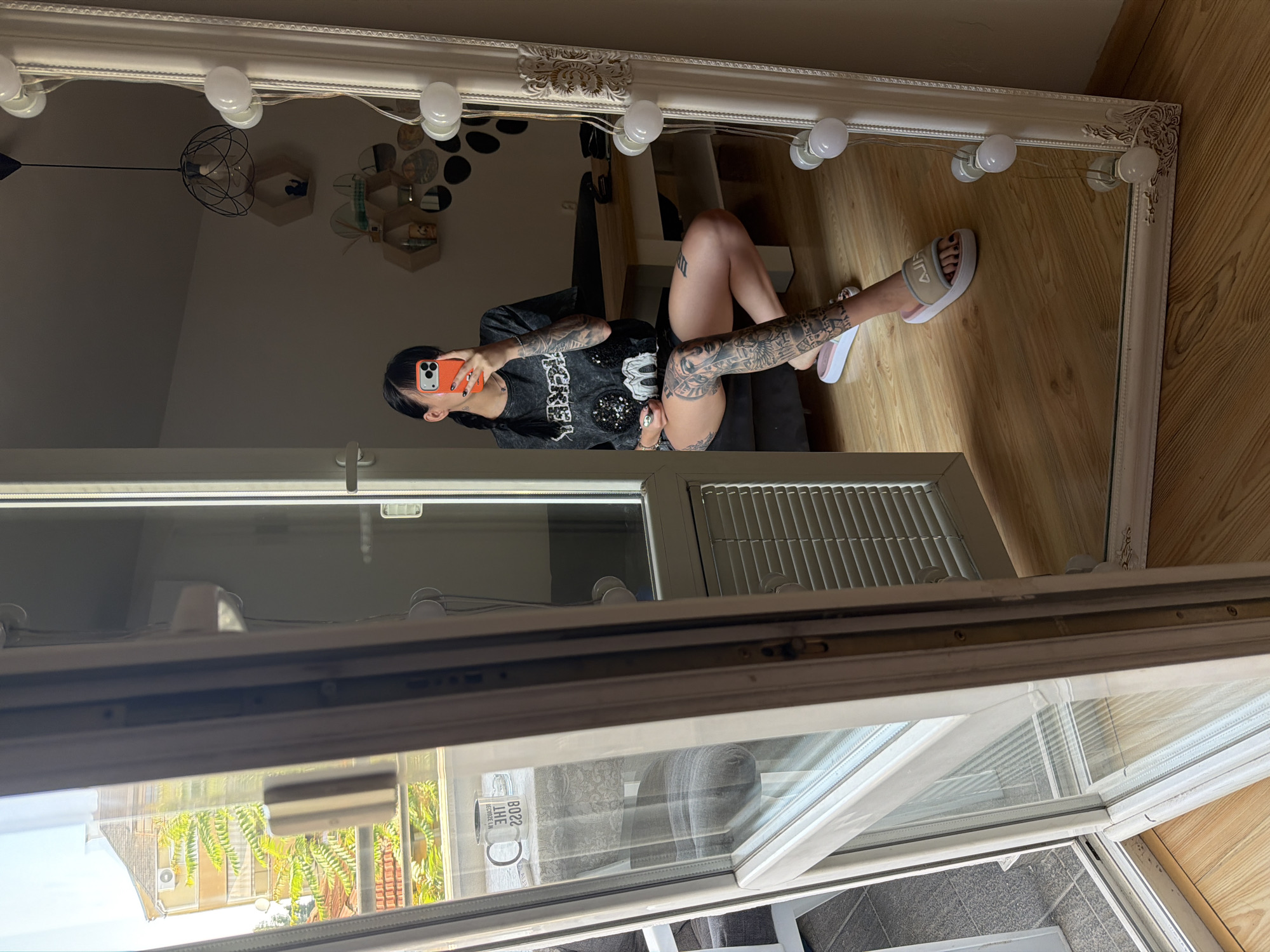 A young adult female is reflected in a large, ornate mirror. She is wearing a dark grey t-shirt with a sequined graphic and dark shorts. Her legs and arms are covered in extensive tattoos. She is holding an orange smartphone in her right hand, taking the selfie. She is wearing white slides with beige accents. The mirror is framed with decorative carvings and illuminated by a string of white lightbulbs. The background visible in the mirror shows a room with wooden floors, a wooden table, a couch, and hexagonal shelves on the wall. Through a doorway and windows on the left side of the image, an exterior view of a building with yellow siding and green trees is visible, suggesting the location is an apartment or house. This scene is set in the city of Plovdiv, Bulgaria.