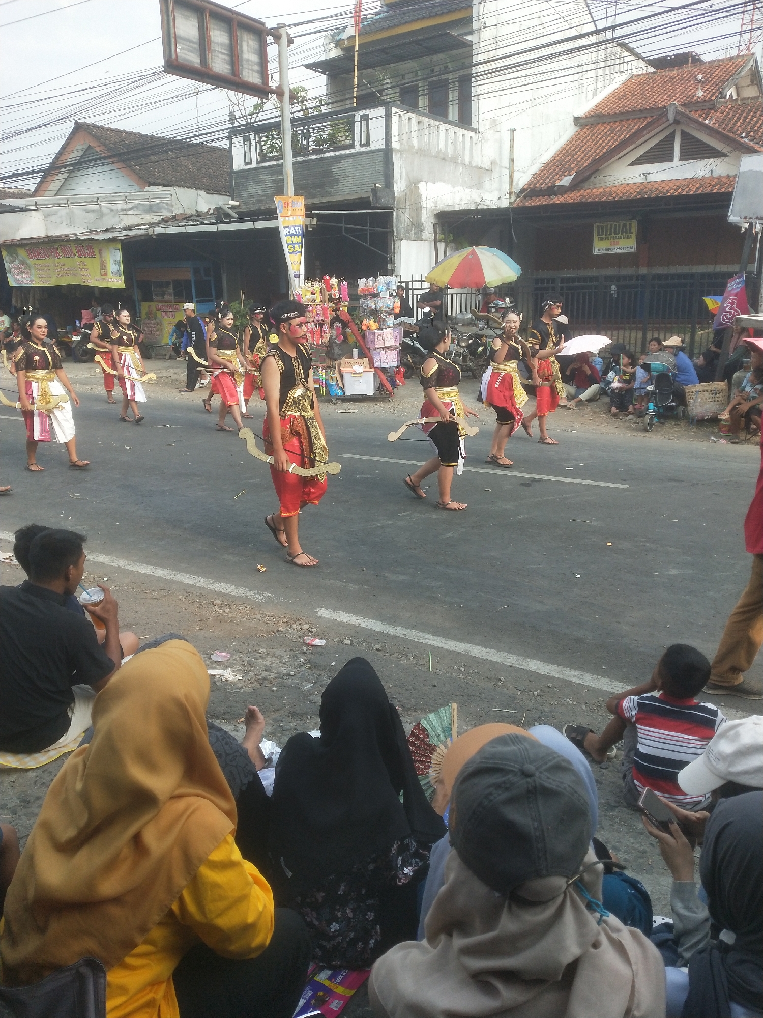 In the city of Boja, Indonesia, a street parade is in progress. Several individuals in traditional attire, some with facial paint, are participating, holding prop weapons resembling bows. They are walking on a paved road lined with spectators seated on the roadside, many of whom are children. Various stalls and buildings are visible in the background, suggesting a lively commercial or residential area. The event appears to be a cultural celebration or festival, with a vibrant atmosphere.