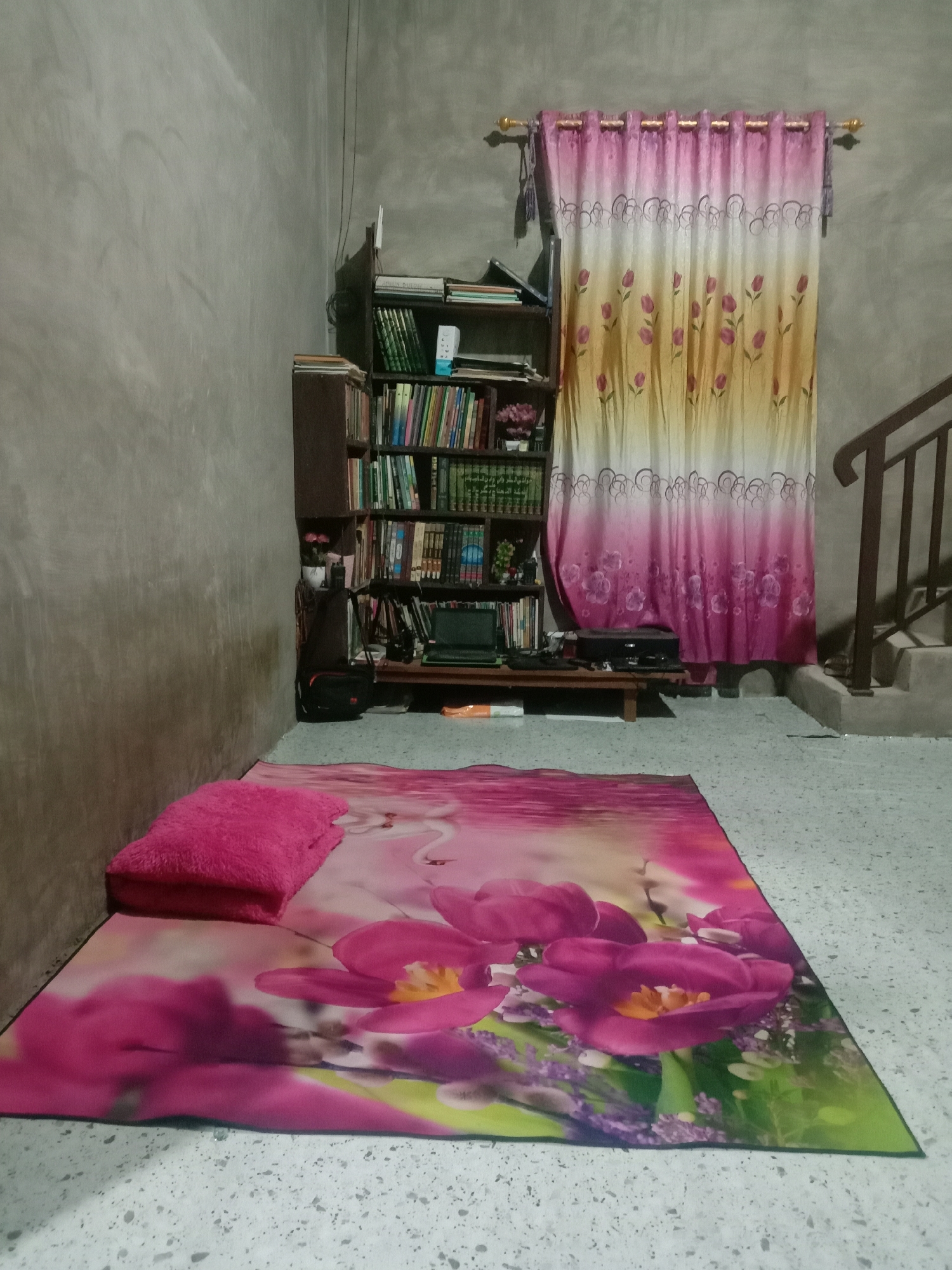 A sparsely furnished room in Lhokjok, Indonesia. The walls are gray, and the floor is a speckled light gray. A floral-patterned mat with a pink pillow sits in the foreground. Behind the mat is a tall, dark-colored bookshelf filled with books. To the right of the bookshelf, a pink and yellow curtain hangs. Stairs are partially visible in the background to the right of the curtain. A low table sits in front of the bookshelf.