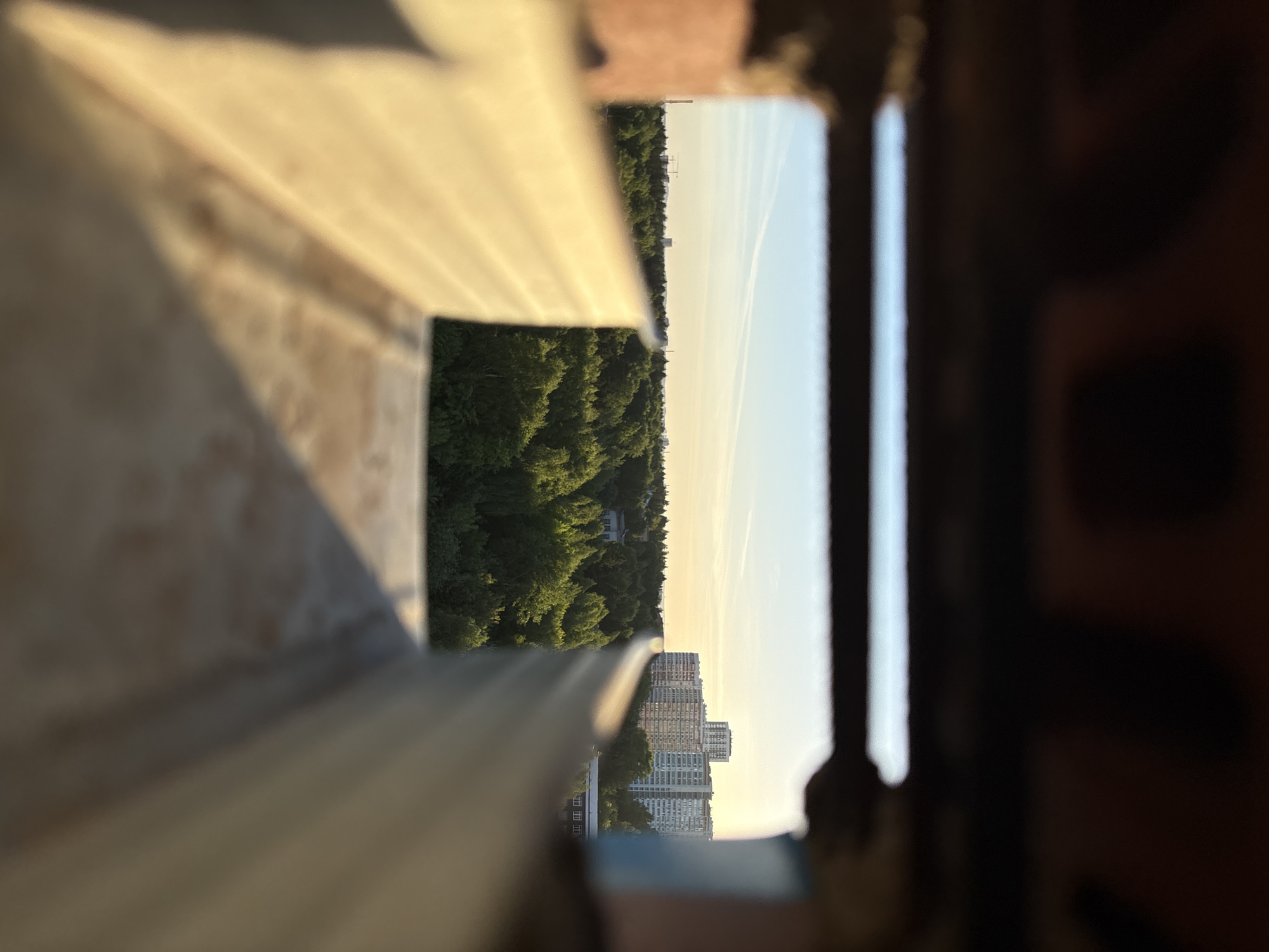 The image is a low-angle, slightly skewed shot taken from an elevated position, possibly a balcony or window. It features a partial view of the surrounding environment of Izhevsk, Russia.
Foreground: The primary focus is a blurred, wooden or concrete structure, likely a railing or ledge, occupying the left and bottom of the frame. The shallow depth of field throws this into soft focus.
Midground: Through the opening created by the structure, there is a view of trees and buildings. A dense forest of green trees is visible, with some trees possibly obscuring objects behind them. Tall apartment buildings are seen in the distance. Above them, the sky is light blue, with what appears to be jet contrails.
Background: On the right side, there is a blurred, dark structure, possibly a part of the building from which the image was taken.
Lighting: The scene is bathed in soft, warm sunlight, suggesting that the photo was taken during the early morning or late afternoon.
Overall Impression: The scene appears serene. The juxtaposition of the natural environment of the forest with the urban landscape of the buildings creates a contrast.