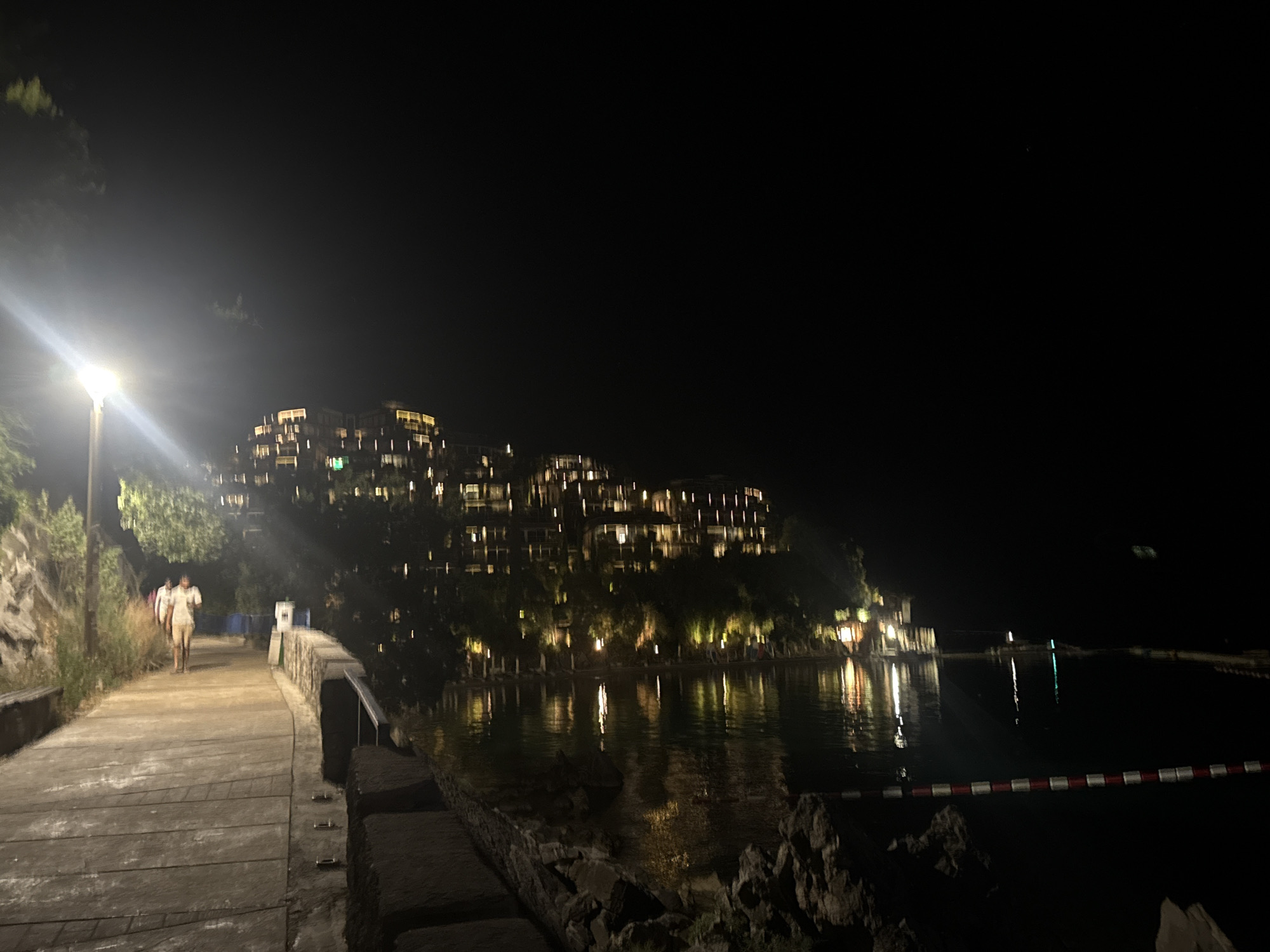The image depicts a waterfront promenade at night in Budva, Montenegro. On the left side of the frame, a pathway illuminated by a streetlamp leads away from the viewer. Two individuals, appearing to be walking away from the camera, are visible on the pathway. Beyond the pathway, lush green foliage and a rocky incline are present. Across the water, a multi-story building complex is illuminated by numerous lights, suggesting it is a hotel or resort. The water itself reflects the lights from the building and the sky, creating a shimmering effect. A red and white rope barrier floats on the surface of the water in the foreground. The overall scene conveys a serene nighttime atmosphere.