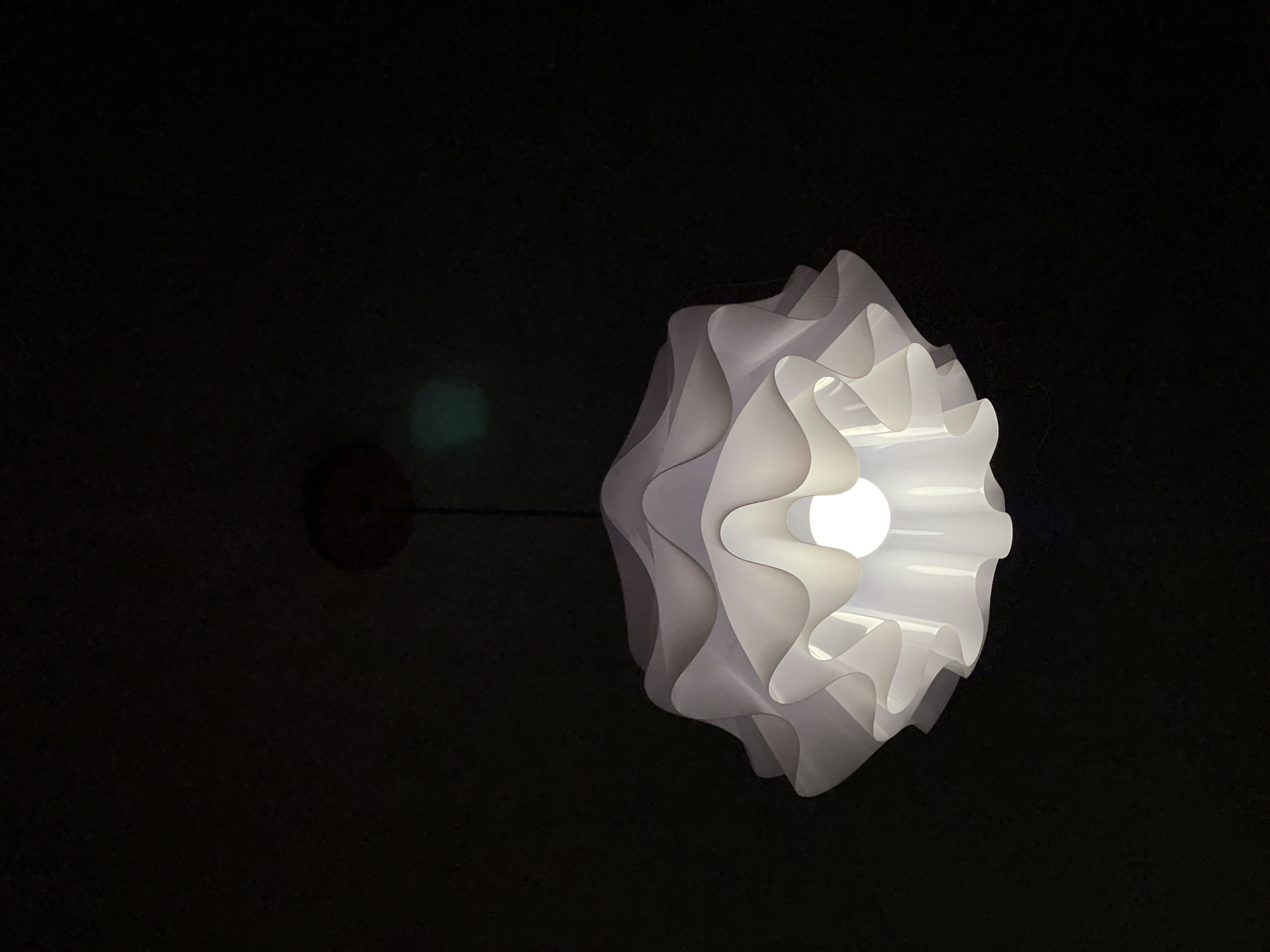The image displays a white, modern-style ceiling lamp illuminated against a completely dark background, suggesting an interior setting at night or in a very dim room. The lamp features a distinctive design with multiple layers of wavy, ruffled, or pleated material, giving it a sculptural, almost organic form. A bright, spherical light bulb is visible within the center of the fixture, acting as the primary light source. The lamp hangs from a small, dark, circular fixture base, which is faintly visible against the dark ceiling to the left. A faint, blurry, greenish-blue light flare or reflection is present in the upper left quadrant of the dark background. There are no people, activities, or discernible text in the image. The focus is solely on the illuminated light fixture and its design.