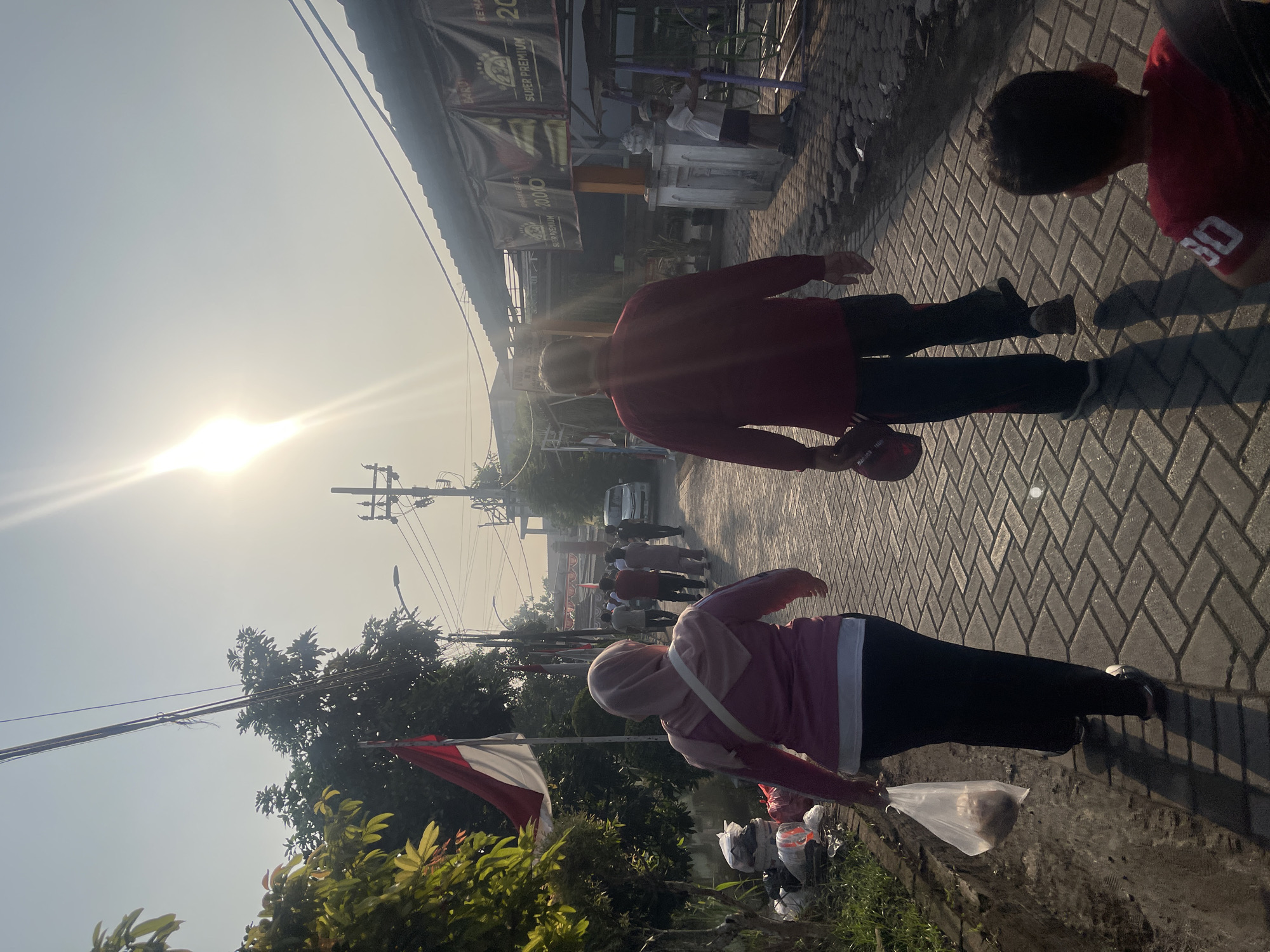 The image captures a street scene in Sugih Waras, Indonesia, under a bright, hazy sun. In the foreground, a woman wearing a pink hijab and a pink jacket walks away from the camera, carrying a white plastic bag with food items in her left hand. She is dressed in black pants and her shadow is cast on the brick-paved street. To her right, a man in a red long-sleeved shirt and dark pants walks ahead, also casting a shadow. He is holding a red cap in his left hand. In the far right foreground, a young child in a red t-shirt is visible from the back.
The street is lined with buildings on the right side, some with dark awnings displaying advertising banners. On the left side, there is vegetation, including a tree with green leaves, and a pile of garbage bags and buckets. Indonesian flags are visible, one large flag on the left and several smaller ones further down the street. Several other people are walking away from the camera in the middle ground, and a white van is parked on the right side of the street. Power lines and utility poles are present overhead. The overall impression is of a normal day in a residential or commercial area.