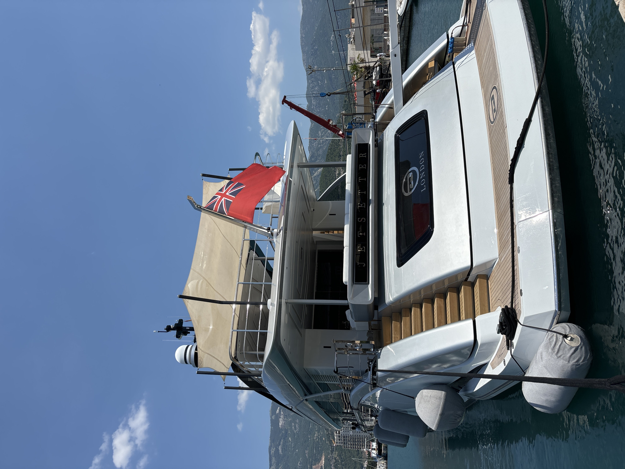 The image shows a large, white yacht docked in a harbor. The yacht has several decks, a Union Jack flag flying from a mast, and the name "JETSETTER" emblazoned on the side. A window on the yacht has the word "LONDON" and a circular logo with an 'E' inside. The yacht is moored with black ropes and has large, grey fenders hanging over its sides. In the background, there are mountains, buildings, and a red crane. The sky is blue with some white clouds, suggesting a clear day. The water around the yacht is a deep teal color. The image is oriented vertically, as if taken from a slightly elevated position looking down at the yacht. The scene appears to be a sunny day in a coastal city, likely Budva, Montenegro, given the context. There are no people prominently visible in the image.