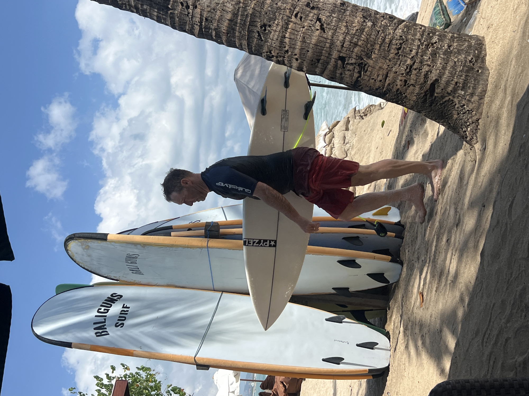 A middle-aged man with short brown hair, wearing a black Quiksilver rash guard and red swim shorts, walks across a sandy beach in Kuta, Indonesia, carrying a white surfboard. He is walking from right to left in the frame. Behind him, several other surfboards stand upright, leaning against a wooden rack. The surfboards are white and orange, with some featuring logos. The man is walking towards the ocean, which is visible in the background, with waves breaking on the shore. The sky is blue with scattered white clouds. A large palm tree trunk is prominently featured on the right side of the frame. The lighting suggests it is daytime, likely late morning or early afternoon. The environment is a tropical beach setting.