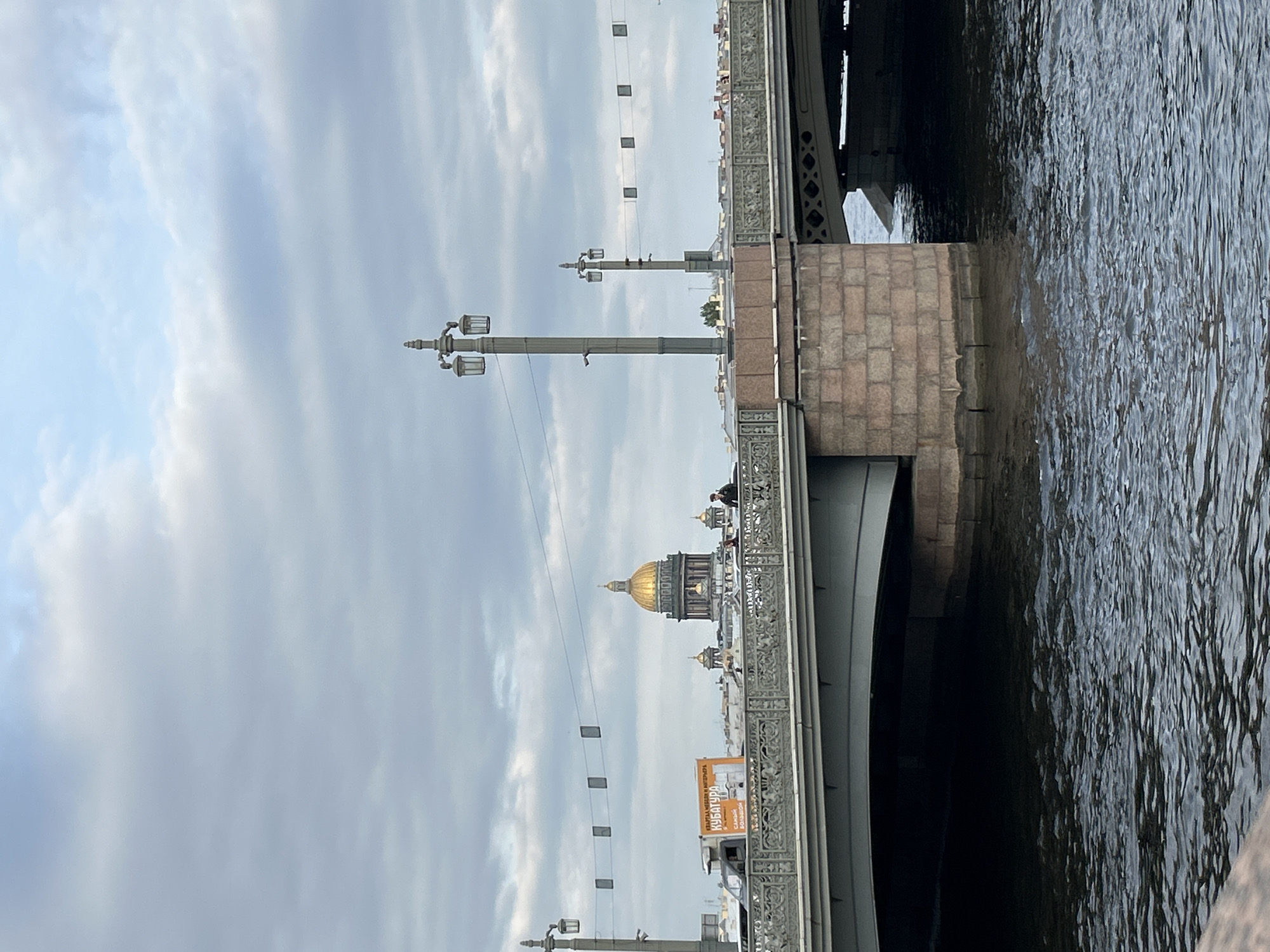 The image shows a view in Saint Petersburg, Russia, featuring the dome of Saint Isaac's Cathedral. The cathedral is viewed from a bridge, identifiable by its ornate railings and supports. The Neva River is visible on the right side of the frame. Streetlights and possibly decorative banners are strung along the bridge. The sky is overcast. There are figures of people on the bridge.