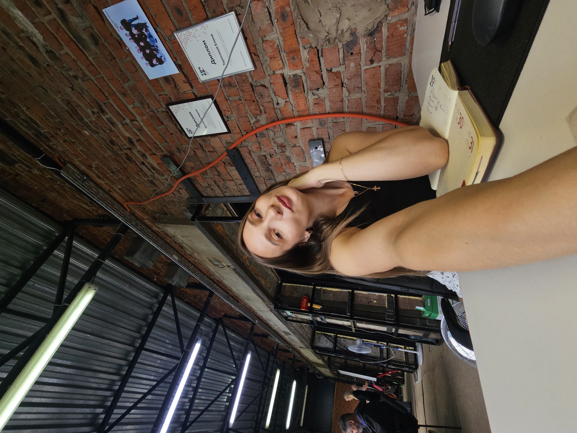 A woman is taking a selfie in a location with a brick ceiling. Mounted on the ceiling are framed documents and a team photo. A notebook is on a table beside her. The environment is industrial-style, with exposed metal structures and fluorescent lights. Further in the background, other people are visible. It is difficult to confirm the location without more information.
