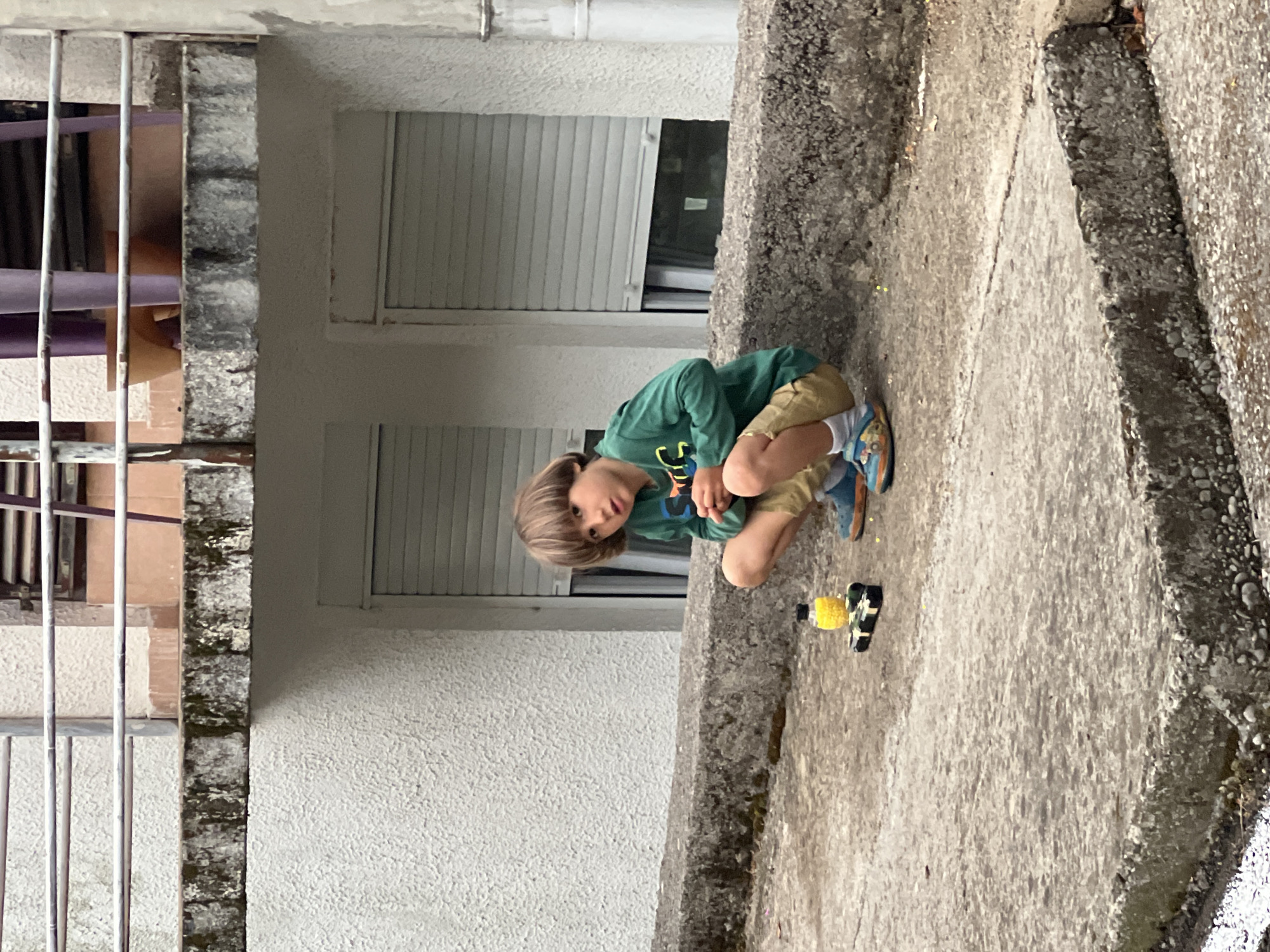 A young boy is crouching on a concrete surface, possibly a low wall or ledge, next to a toy car. The boy is wearing a green long-sleeved shirt and khaki shorts. He is looking towards the camera. A building with white walls and closed window shutters is in the background. A metal railing is visible on the side of the building. The concrete surface is textured with some signs of wear and possible moss growth. Based on the architectural style and general appearance, the location may be Budva, Montenegro.