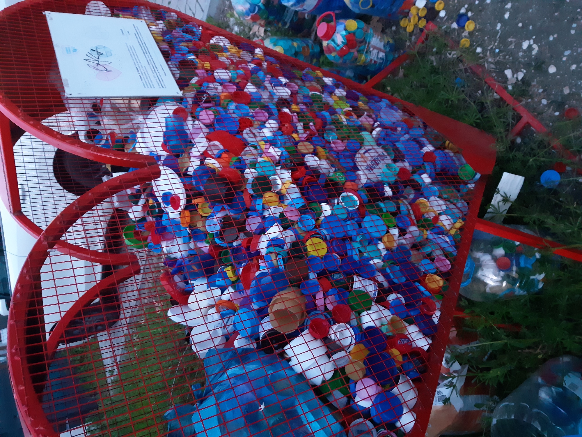 The image depicts a red metal structure filled with an assortment of plastic bottle caps, predominantly blue, red, white, and yellow. The structure appears to be a decorative collection point for recycling, judging by the accumulation of bottle caps within it. There is a sign placed on the structure. The location is Plovdiv, Bulgaria. There is vegetation around the structure. Also there are some water bottles lying around. The light condition looks like it's sunny outside.