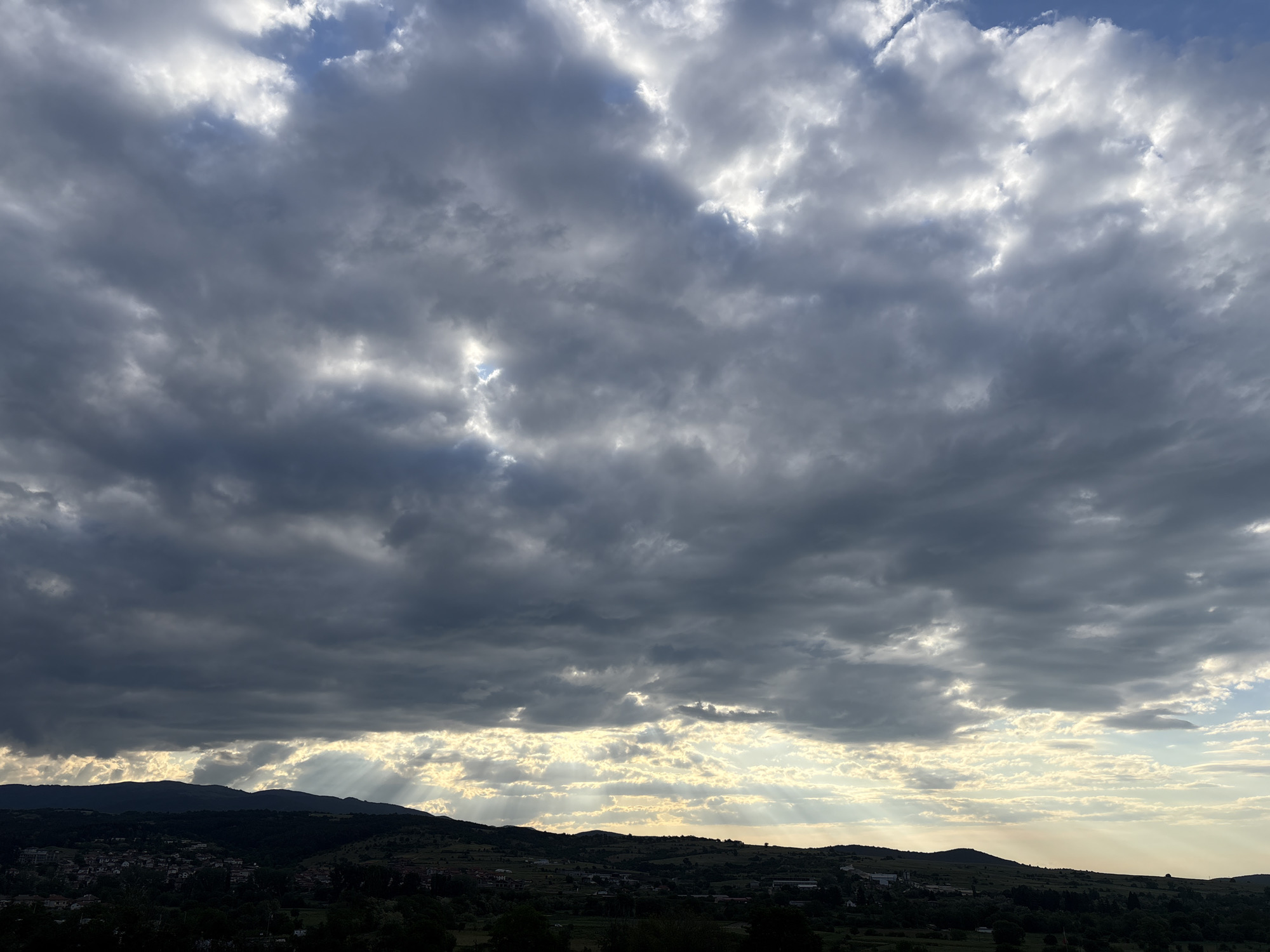 The image depicts a landscape in Strelcha, Bulgaria. In the foreground, there is a village nestled among rolling hills and trees. In the background, there is a dramatic sky filled with dark, heavy clouds. Sunlight streams through gaps in the clouds, creating sunbeams that illuminate parts of the landscape below.