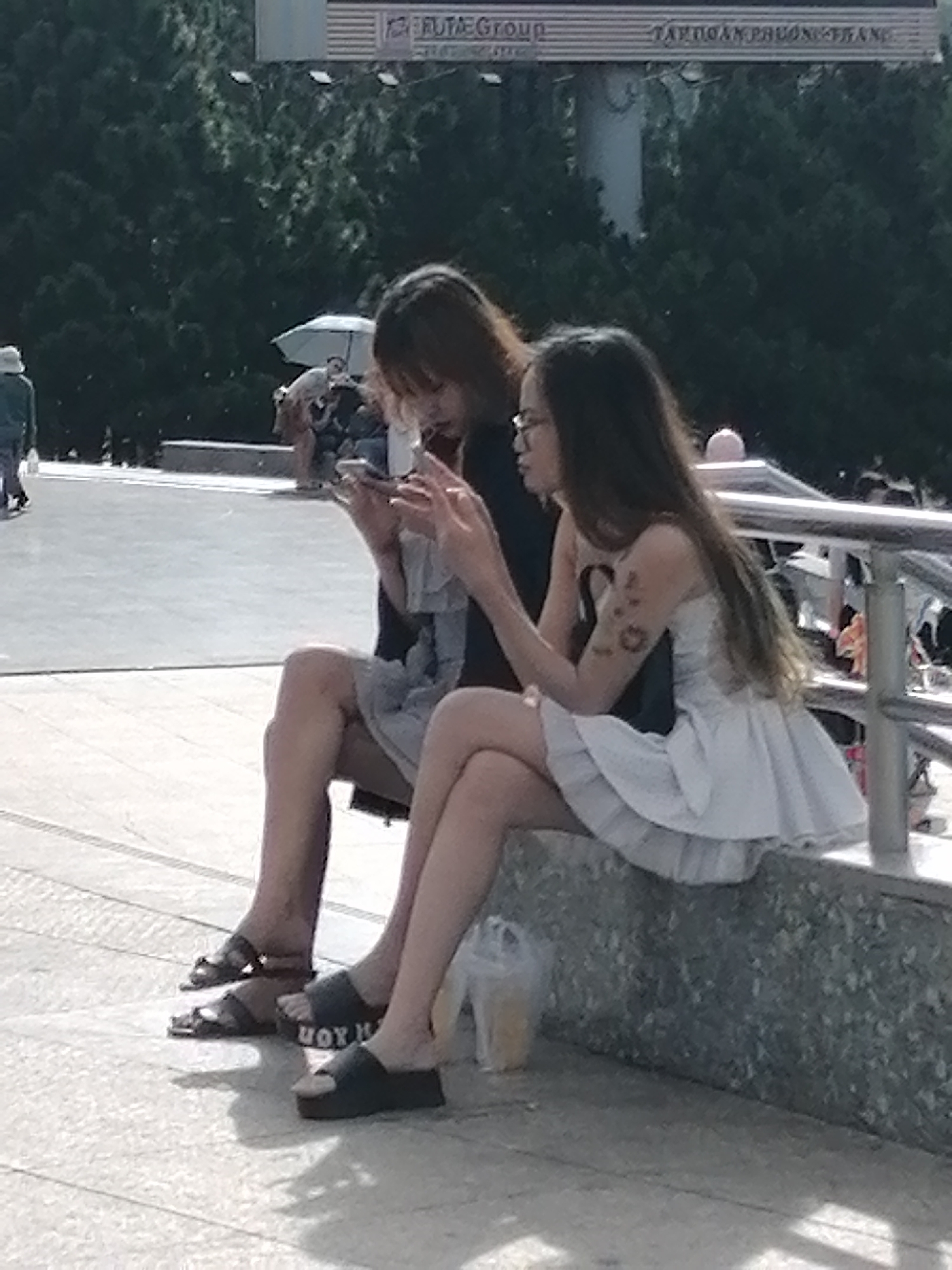 Two young Vietnamese women are sitting on a concrete ledge outdoors, looking at their mobile phones. The woman on the left has long, dark brown hair and is wearing a black tank top and grey shorts. She has dark sandals on. The woman on the right has long, light brown hair and is wearing glasses and a white strapless dress with a ruffled hem. She has dark, slip-on sandals on. A plastic cup with a yellow drink is on the ledge next to the woman on the right. In the background, there are trees, a metal railing, and a billboard. The scene appears to be in Da Lat, Vietnam, likely a public area near a tourist attraction, given the presence of a billboard and the general atmosphere.