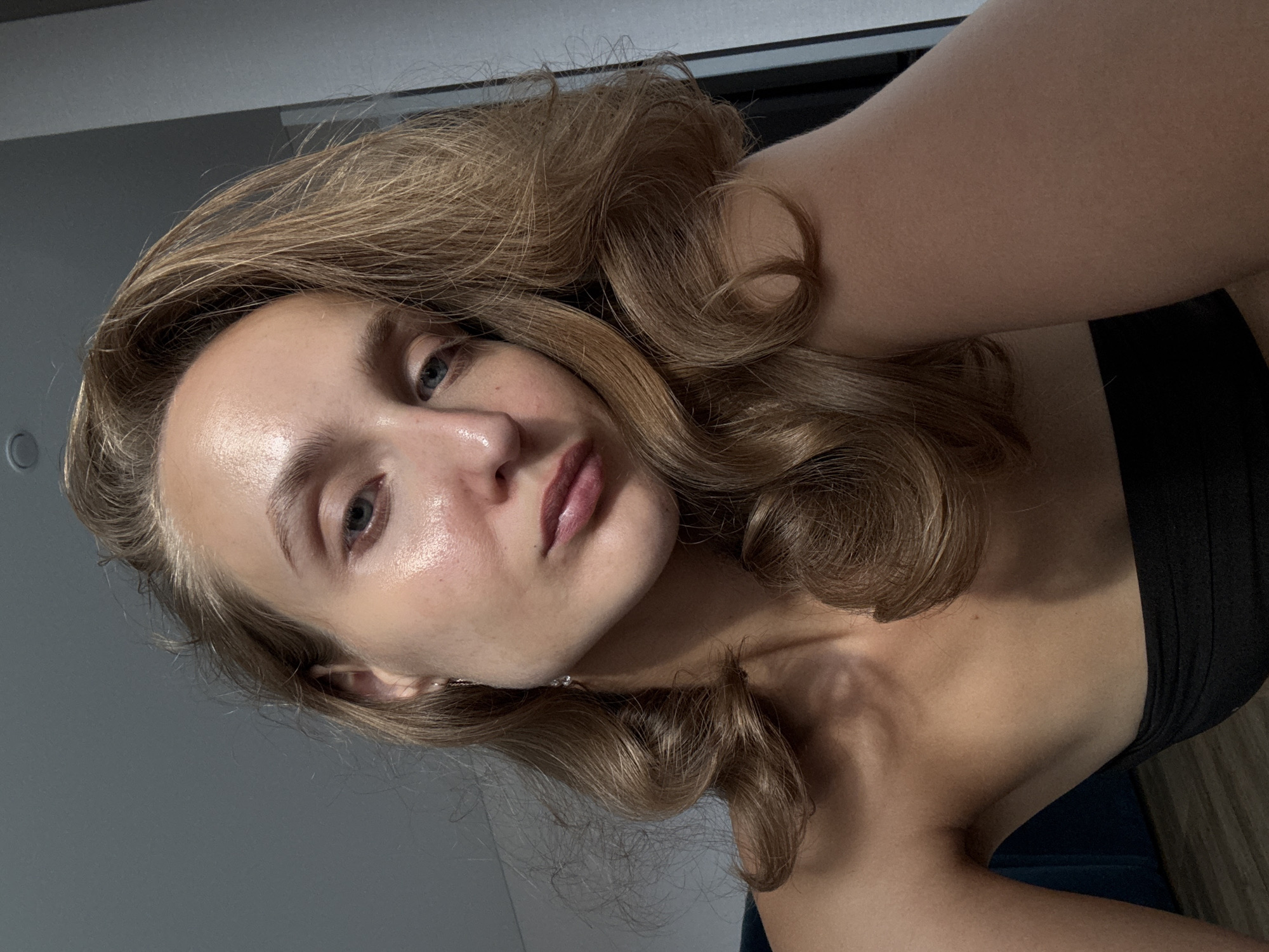 A selfie is taken by a young woman with light skin and blonde hair, styled in loose waves. She is wearing a black strapless top. Her left eye is blue, and her lips are slightly parted, showing a pout. The background is out of focus, but suggests an indoor setting with grey walls and a wooden floor. The lighting is soft, highlighting the sheen on her skin. There is no identifiable context for the image to determine location within the city of Podshivalovo, Russia, or any specific activity.