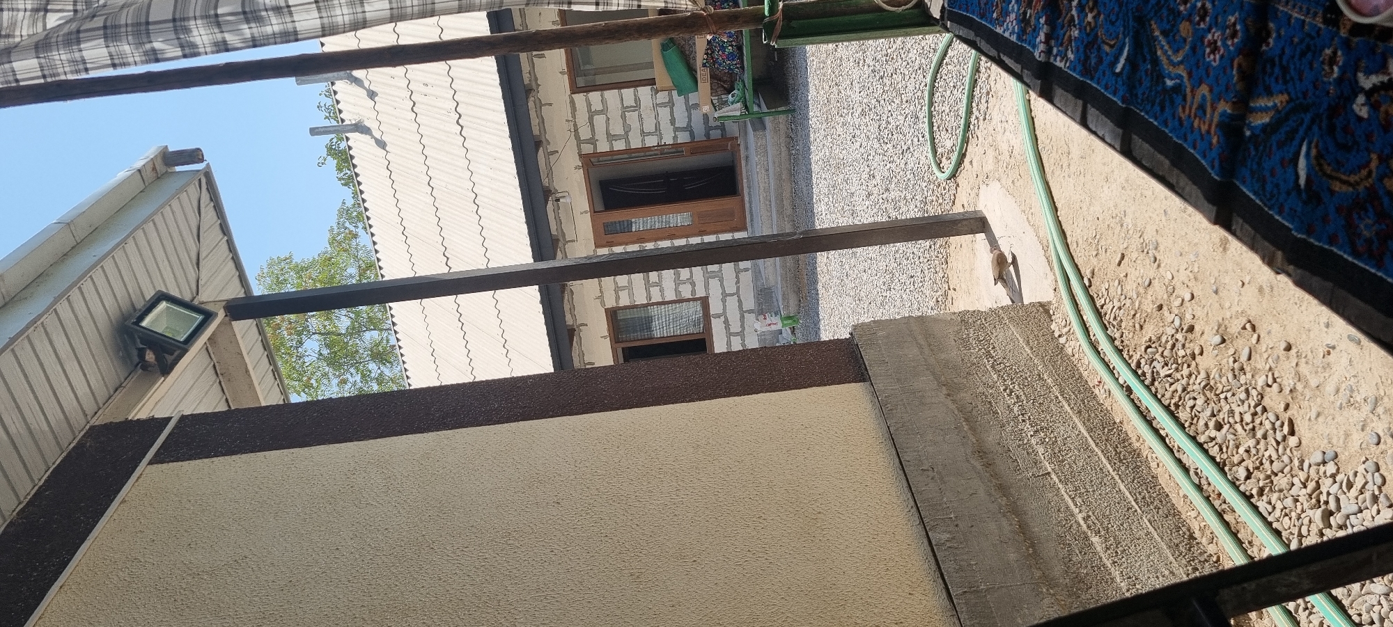The image captures an outdoor scene in Ҷомӣ, Tajikistan, on a bright, sunny day. The foreground is dominated by a textured, light-colored wall and a concrete foundation, with two green hoses lying on the ground. In the middle ground, a building constructed of light-colored bricks is visible, featuring wooden doors and windows. A gravel-covered area surrounds the building. To the right, a green metal bench is placed near some plants. The background shows a light-colored, corrugated roofline and trees under a clear blue sky. A bird is visible walking on the ground to the right of the concrete foundation. A light fixture is mounted under the overhang of a metallic roof to the left.