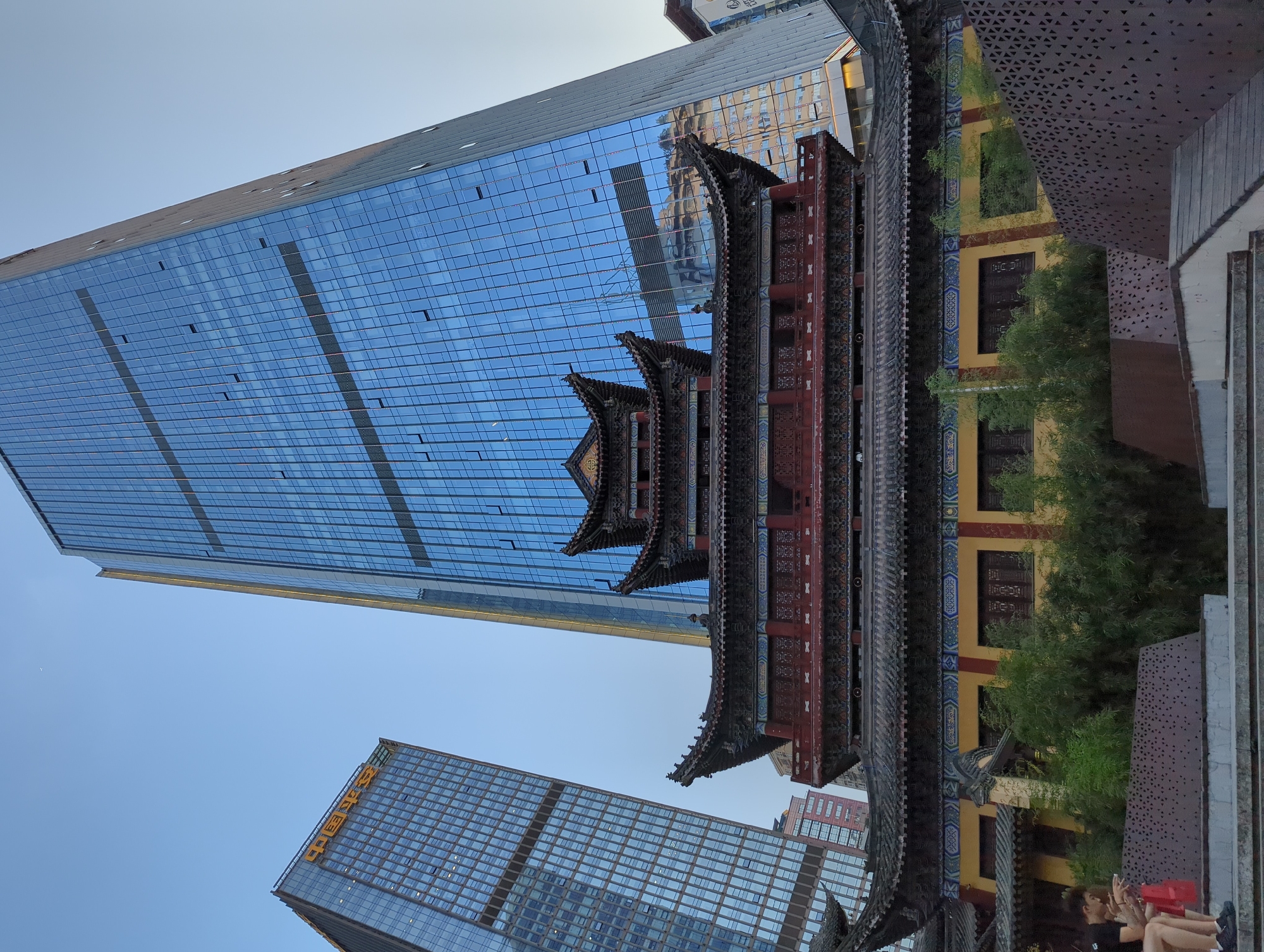 The image captures a juxtaposition of traditional Chinese architecture and modern skyscrapers in Yuzhong District, China. In the foreground, a multi-tiered, ornate traditional building with a dark tiled roof and red and yellow accents stands prominently. The building features intricate decorative elements along its eaves and walls, with dark wooden windows visible. Green bamboo plants are growing around the base of this traditional structure.
Behind the traditional building, two modern skyscrapers dominate the skyline. The one on the right is significantly taller and features a sleek, blue glass facade with a slightly angled design, reflecting the sky and surrounding buildings. A smaller, lighter-colored skyscraper is visible to the left, also with a glass facade and featuring some Chinese characters, likely a company logo, in yellow at its top.
In the lower left corner of the image, two individuals are seated on a concrete ledge. One is wearing a black t-shirt and shorts, while the other is wearing a red shirt and shorts. Both appear to be looking at their phones, with one holding a red bag. A textured, dark metal railing with a geometric pattern runs along the edge of the ledge. Steps leading up to the area are visible in the foreground, made of concrete and wood. The sky is clear and a pale blue, indicating it is likely late afternoon or early evening.