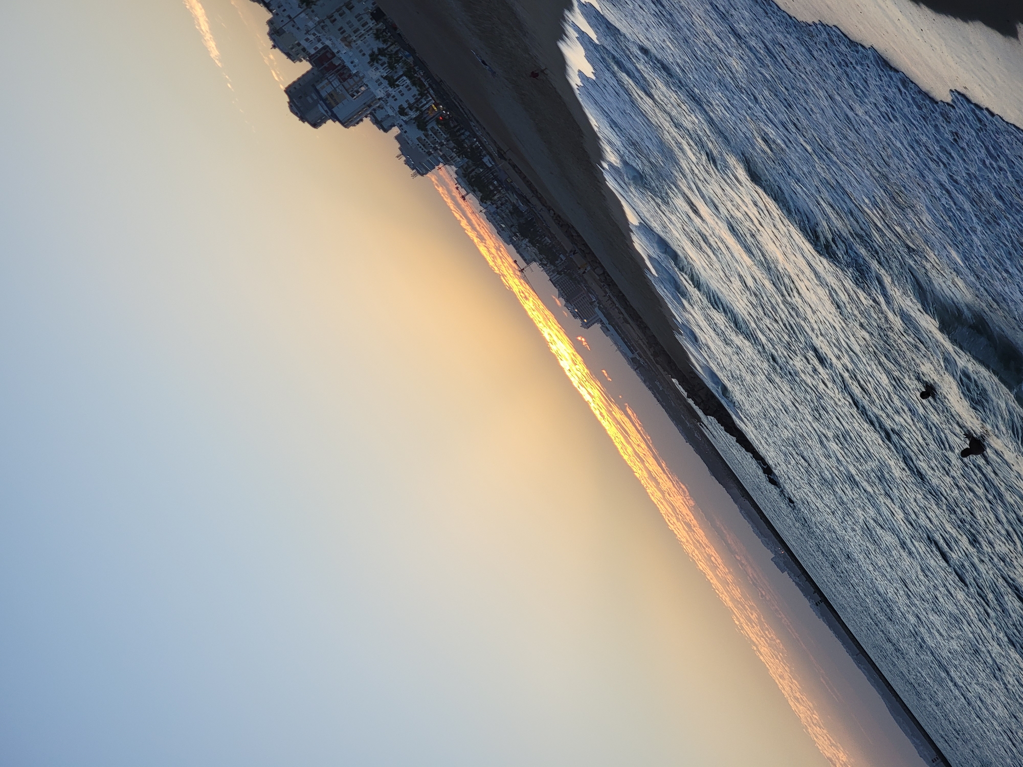 The image, taken in Quarteira, Portugal, depicts a beach scene at dusk. The photograph is oriented diagonally, with the sky occupying the upper left portion and the sea and beach occupying the lower right. The sky transitions from a pale blue at the top to a warm orange and yellow hue near the horizon, indicative of sunset. Wisps of clouds are visible in the sky, catching the last rays of sunlight.
Along the coastline, a strip of buildings forms a prominent feature. These appear to be multi-story structures, some of which resemble apartment buildings or hotels, with white and blue facades. Palm trees are interspersed among the buildings, suggesting a Mediterranean or coastal climate. A crane is also visible amongst the buildings, indicating ongoing construction.
The beach itself is sandy, with a rocky breakwater extending into the sea. The sea is calm, with gentle waves lapping at the shore. Several small, dark figures are visible in the water, which appear to be birds or possibly very small boats. One figure on the beach, to the right of the frame, appears to be a person walking. The overall atmosphere is serene and tranquil, capturing a moment of natural beauty at the end of the day.
