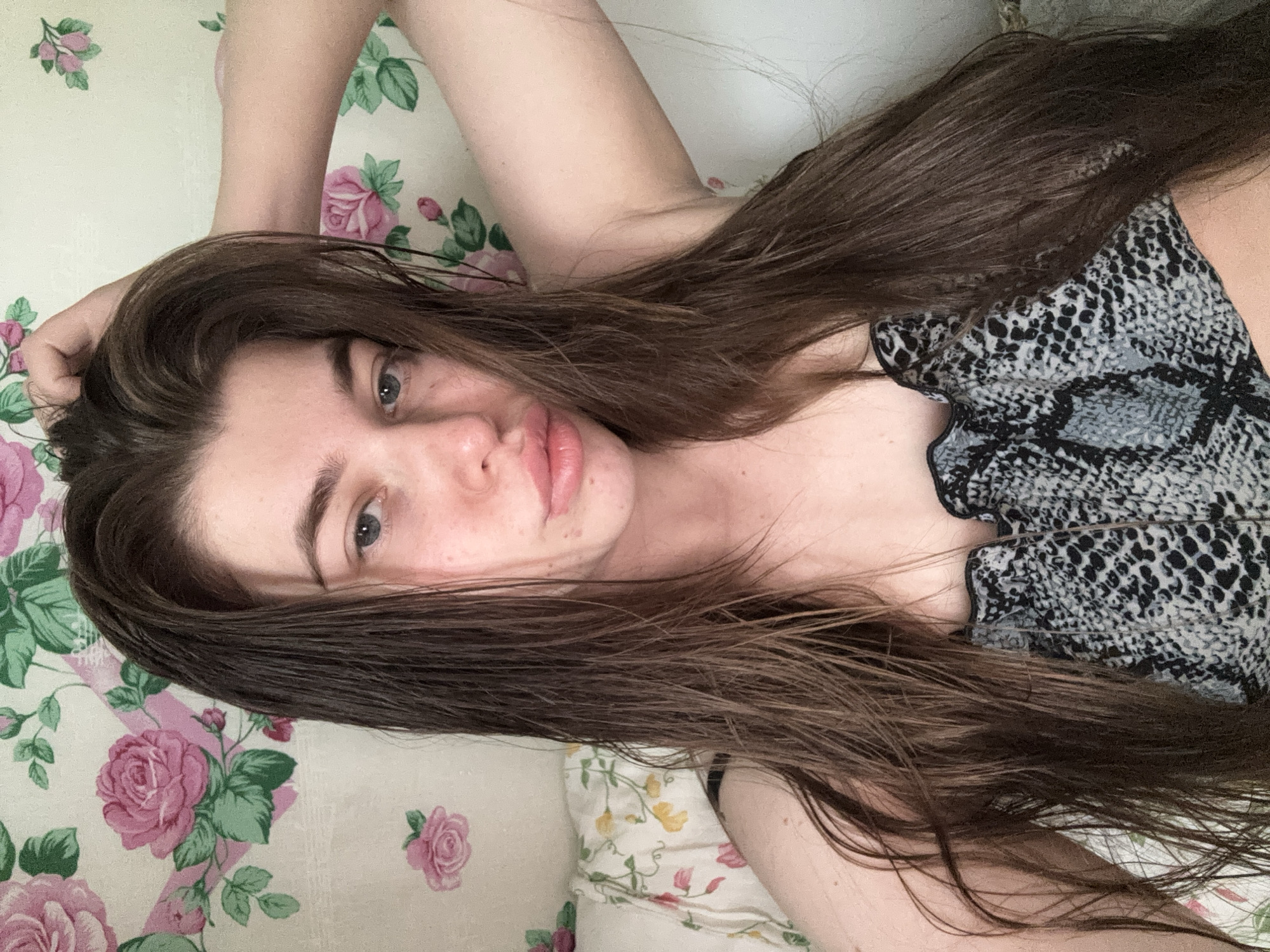 A woman is lying on a floral patterned surface. She has long brown hair, blue eyes, and is wearing a snake print top. Her right arm is raised, and her hand is touching her hair. The background has floral patterns. The location is not identifiable from this image.