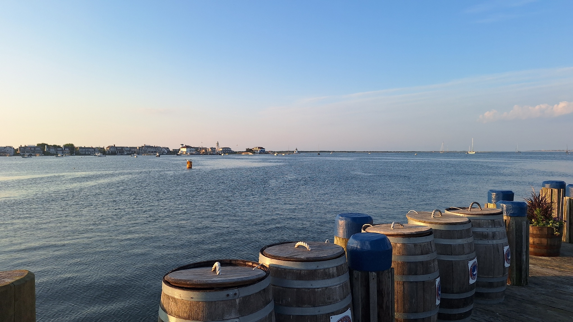 The image depicts a waterfront scene in Nantucket, Massachusetts, United States of America, likely during late afternoon or early evening due to the warm lighting. In the foreground, several wooden barrels with rope handles on their lids are positioned on a wooden dock. Some barrels are topped with blue covers. To the right, there are wooden posts with similar blue covers, and a planter with red foliage. The middle ground features a calm body of water, possibly a harbor or bay, with several boats and yachts sailing in the distance. On the far shore, a line of houses and buildings, characteristic of Nantucket architecture, stretches along the coastline, with a prominent lighthouse visible. The sky is clear and blue, transitioning to a softer hue near the horizon with a few wispy clouds. The overall atmosphere is tranquil and picturesque, capturing a classic New England coastal view.