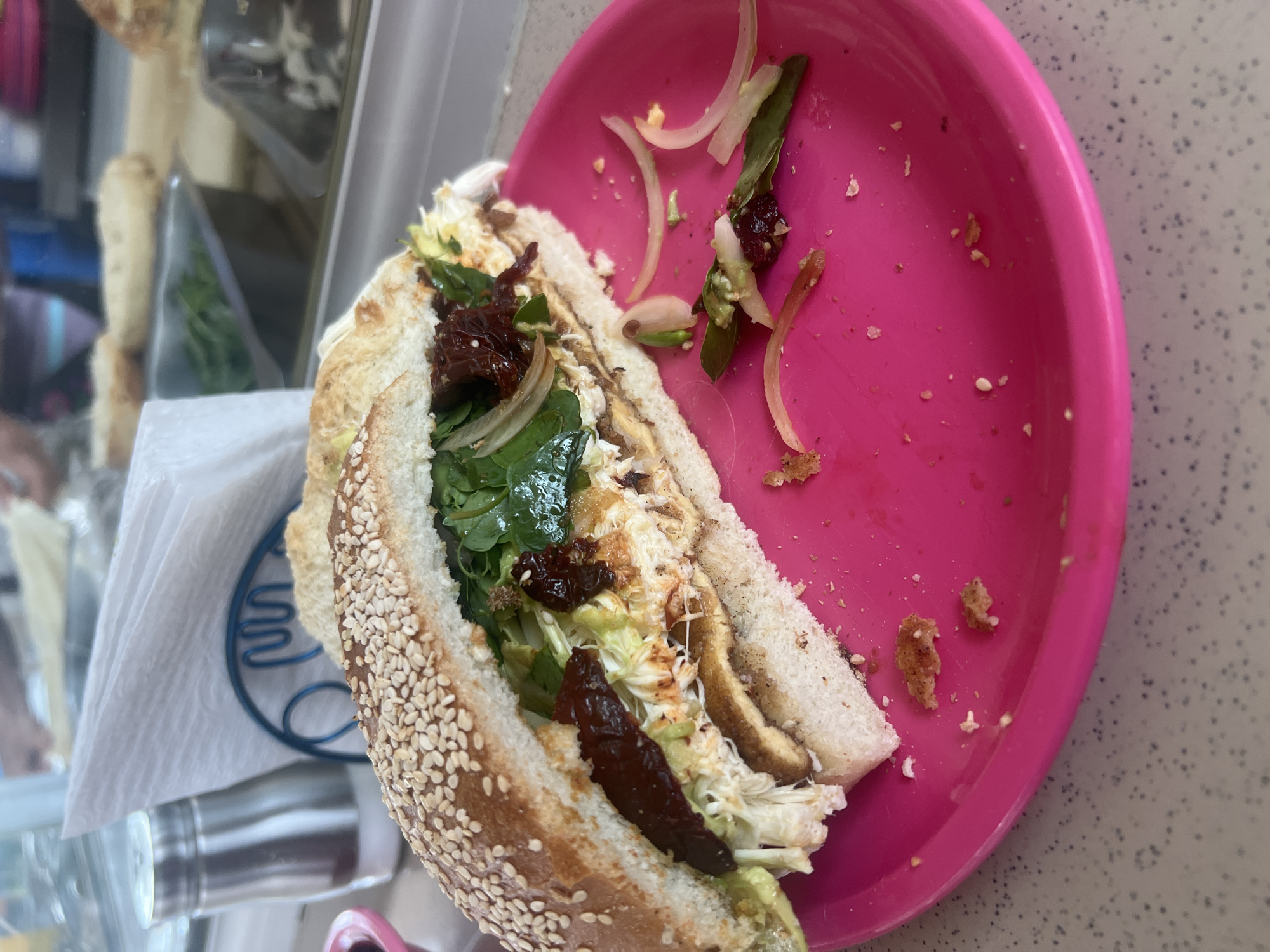 The image shows a partially eaten sandwich on a pink plastic plate. The sandwich is made with sesame seed bread and appears to contain lettuce, spinach, shredded cabbage, dried tomatoes, and onion. The plate also has a few scraps of onion and leafy greens. The sandwich is placed on a white napkin that is on top of a table next to a metallic object. The scene is likely in Puebla, Mexico, because of the style of the sandwich and the plate.