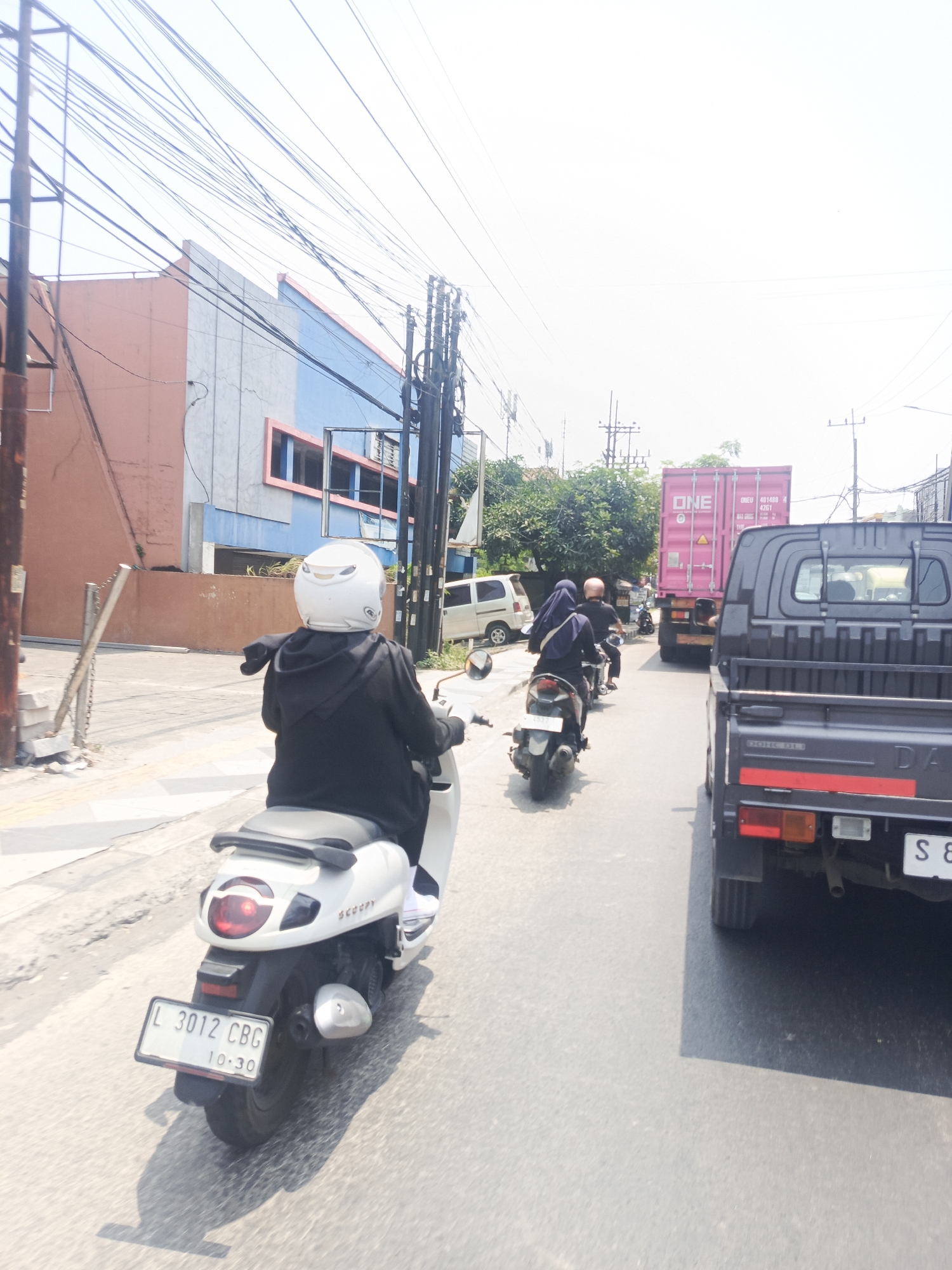 The image captures a street scene in Kepuh Kiriman, Indonesia, on a bright, sunny day. In the foreground, a person wearing a black hoodie and a white helmet rides a white scooter with a license plate reading "L 3012 CBG 10-30". The scooter is in motion, as indicated by the motion blur of the road and the rider's shadow.
Behind the scooter, several other motorcycles are visible. The first one carries two individuals, one wearing a dark blue hijab and the other a dark shirt. Further down the road, another motorcycle is partly obscured.
The street is lined with buildings, including a multi-story structure with a pink and blue facade on the left. Overhead, a complex network of power lines crisscrosses the sky. In the distance, a large pink shipping container, labeled "ONE", is mounted on a truck. To the right, the rear of a dark-colored pickup truck is prominent, with its license plate partially visible. The road surface is asphalt, and sunlight casts distinct shadows.