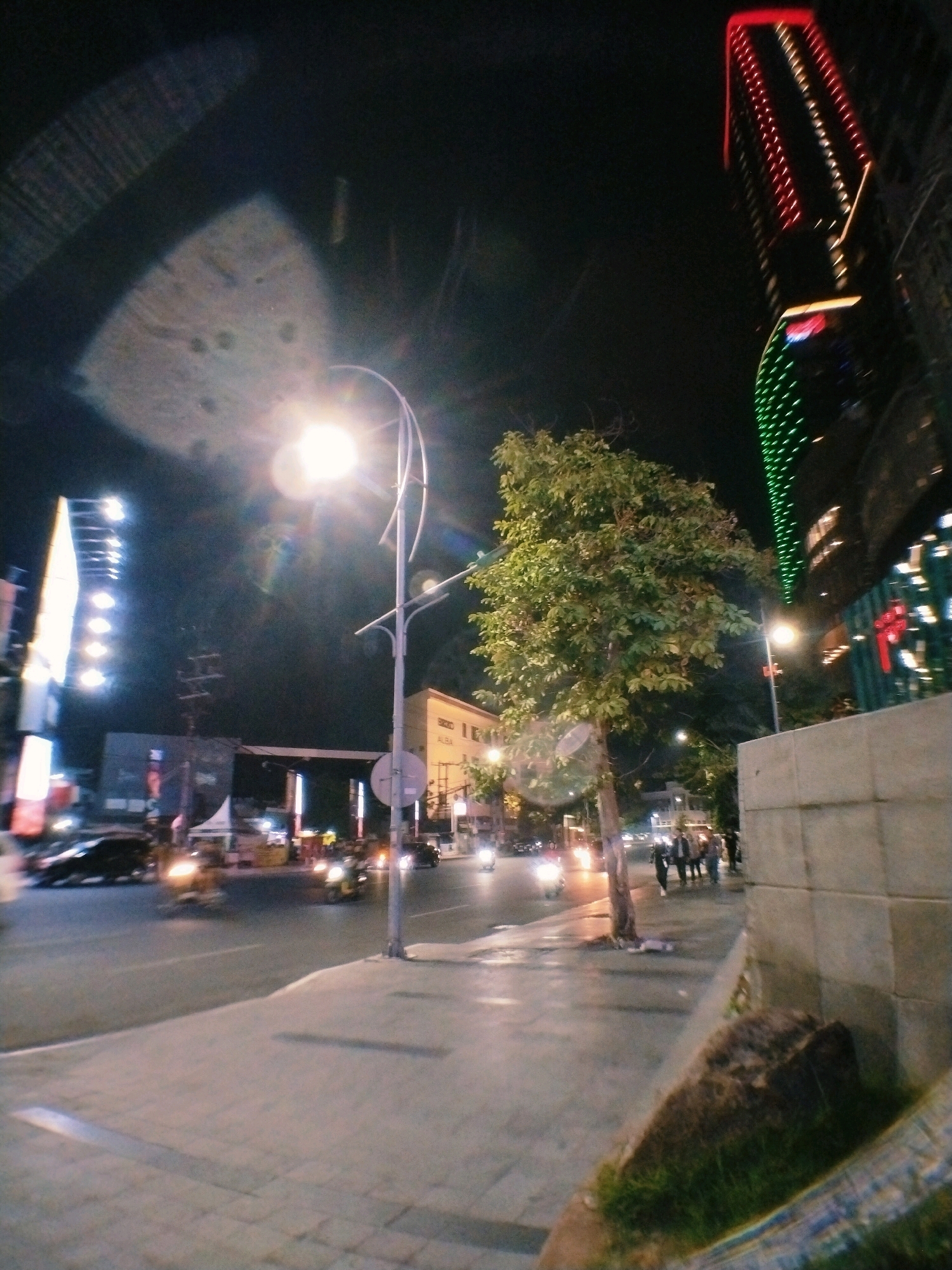 The image depicts a street scene at night in Surabaya, Indonesia. A tall, modern building with red and green lights is visible on the right side of the frame. Across the street, a large billboard with bright lights illuminates the scene. A street lamp with a bright light source is prominent in the foreground, casting a glow on the sidewalk and the street. Several vehicles, including cars and motorcycles, are moving on the road, their headlights and taillights creating streaks of light. A tree with lush green foliage stands near the sidewalk. A group of people are walking on the sidewalk in the background. The street is paved with asphalt, and the sidewalk is made of interlocking bricks. The overall atmosphere is that of a busy urban environment at night.