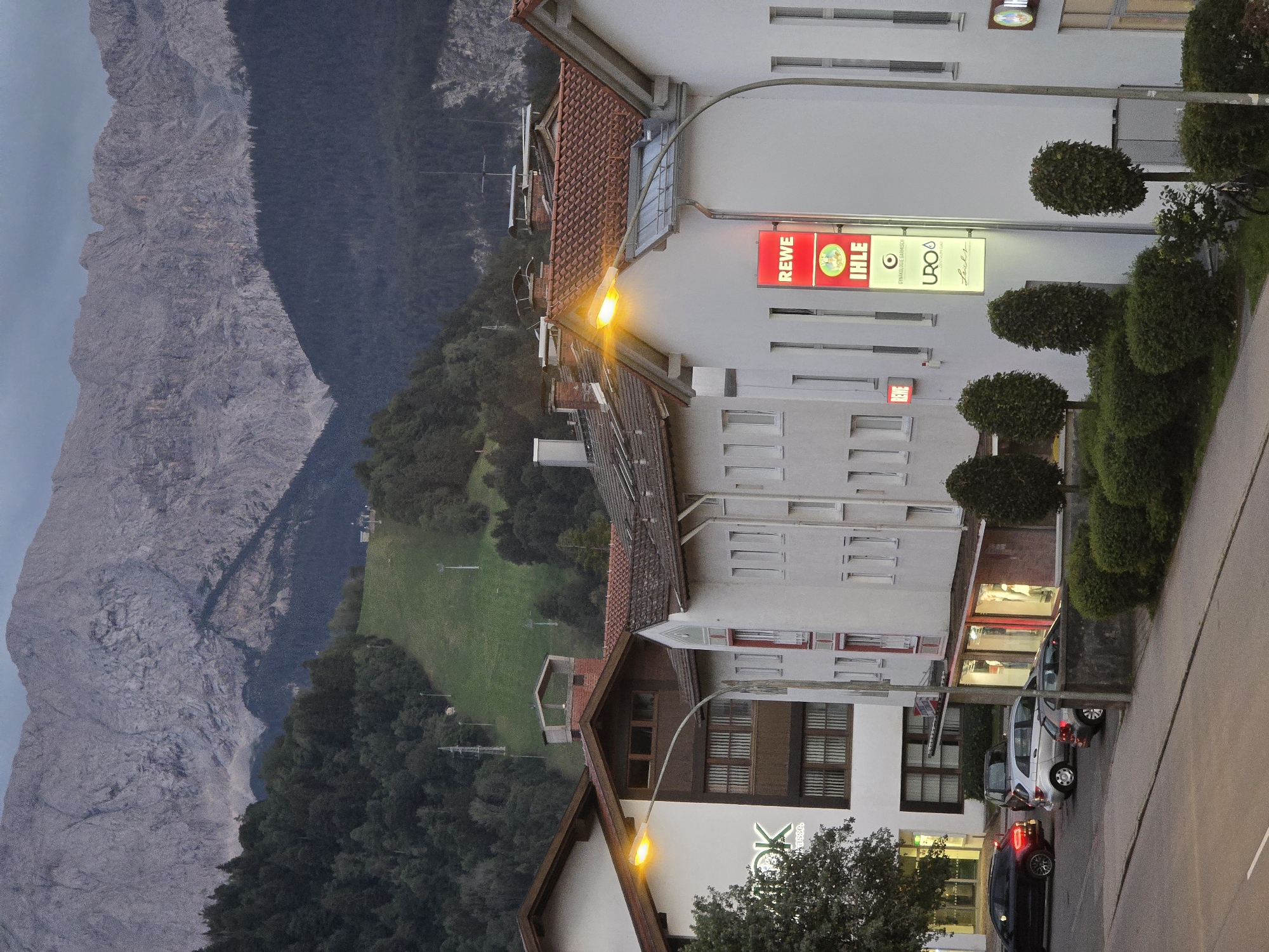 In Garmisch-Partenkirchen, Germany, a street-level view captures a twilight scene dominated by a majestic mountain range in the background. The lower slopes of the mountains are covered in a dense, dark forest, transitioning to a grassy ski slope further down. Several ski lift poles are visible on the slope. In the mid-ground, a row of white buildings with red-tiled roofs lines the street. One building prominently features a bright orange streetlamp casting a warm glow. Signs on the buildings indicate businesses, including "OK" and "REWE IHLE." The street in the foreground is lined with parked cars, including a black sedan and a silver hatchback. Greenery, including well-maintained bushes and trees, borders the sidewalk. The sky is overcast, suggesting the late hour and the approaching evening.