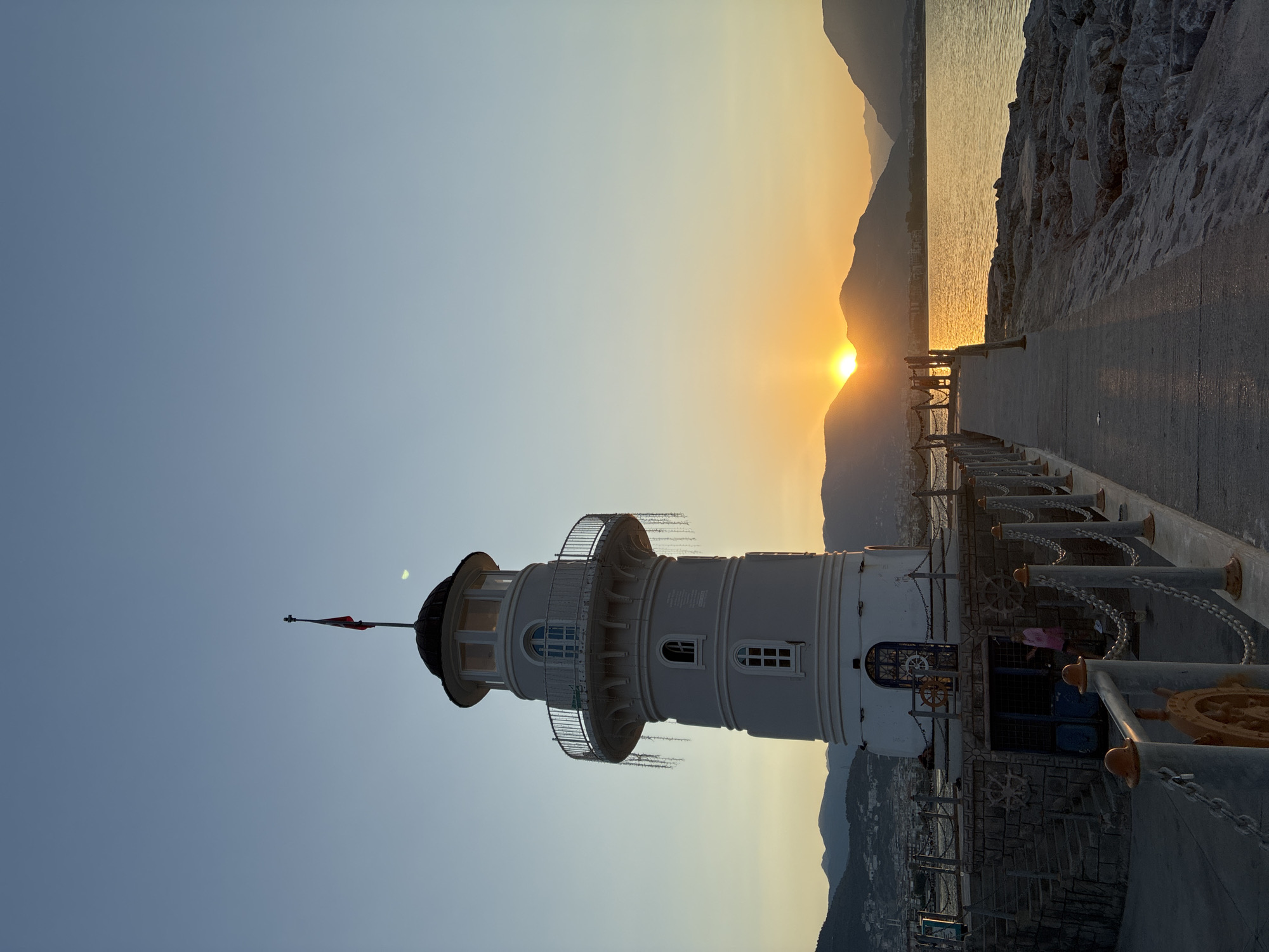 The image depicts a lighthouse at sunset in Alanya, Turkey. The lighthouse, a white, cylindrical structure with a dark conical roof and a flag at its apex, stands on a stone pier. It has several windows, including arched ones on the upper levels and rectangular ones below. Decorative elements, possibly fairy lights, hang from the balcony.
A concrete walkway extends from the lighthouse, lined with a metal chain railing supported by posts. The pier itself is constructed of stone. In the background, the sun is setting behind a range of mountains, casting a warm, golden glow across the sky and the calm sea. The coastline of Alanya is visible in the distance, with buildings and hills dotting the landscape.
A few people are present. Two individuals, one wearing a pink garment, are near the base of the lighthouse, close to an entrance. Another person is further down the walkway. The sea is calm, reflecting the colors of the sunset. The overall atmosphere is serene and picturesque.