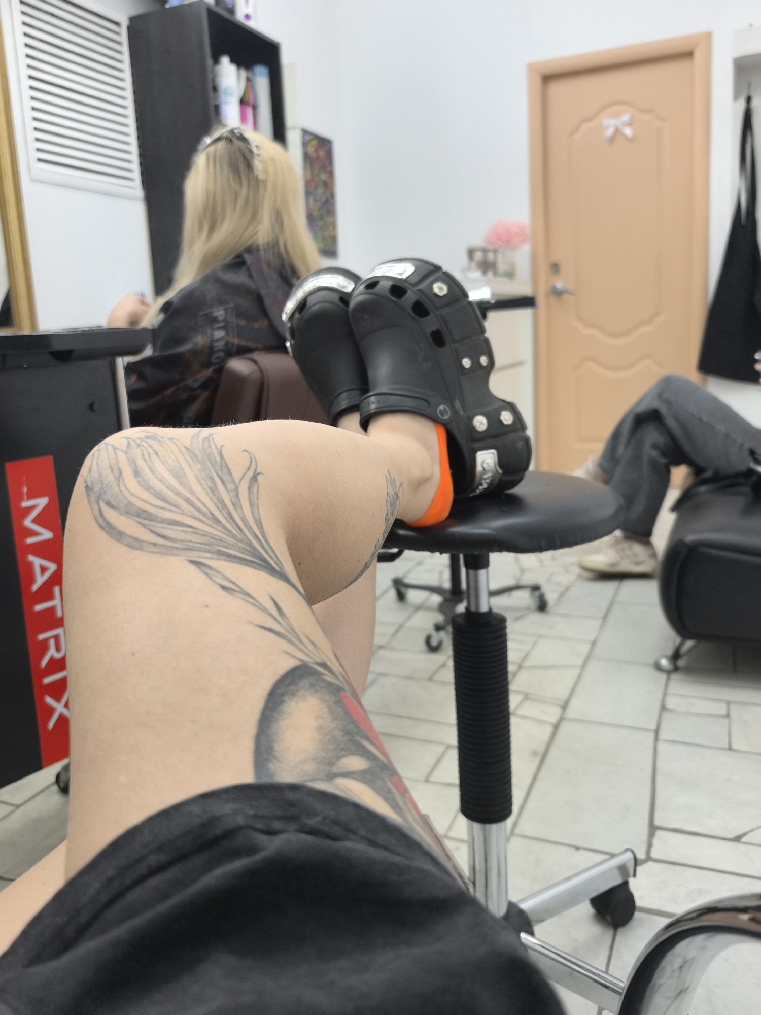 The image depicts a low-angle view of a person's legs and feet, wearing black Crocs with orange accents, resting on a black swivel chair in what appears to be a salon or barbershop. A large floral tattoo is visible on the person's left leg. In the background, a woman with blonde hair is seated, facing away from the camera. Other elements in the background include shelves with bottles, a beige door with a bow, and tiled flooring. The overall setting suggests an indoor commercial establishment. The location is not specified in the image itself or its metadata.