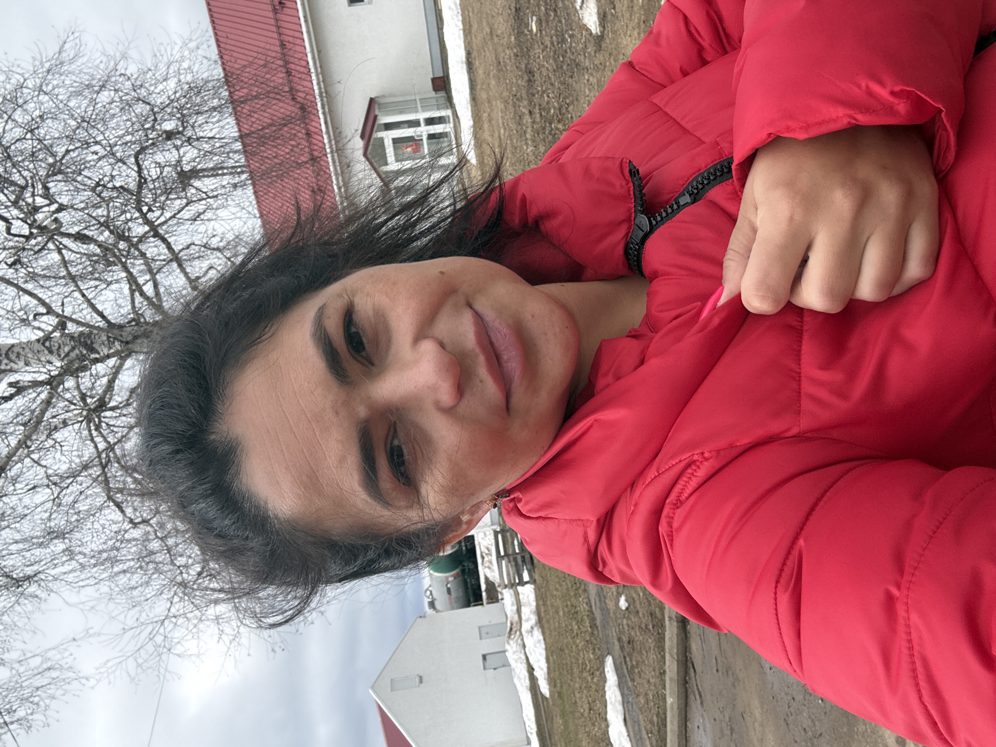 A woman is seen in a selfie style shot. She is wearing a red puffer jacket. The background includes a birch tree with bare branches, a white building with a red roof, and patches of snow on the ground. The location is consistent with an outdoor setting in Kauchuk Stantsiya, Russia.