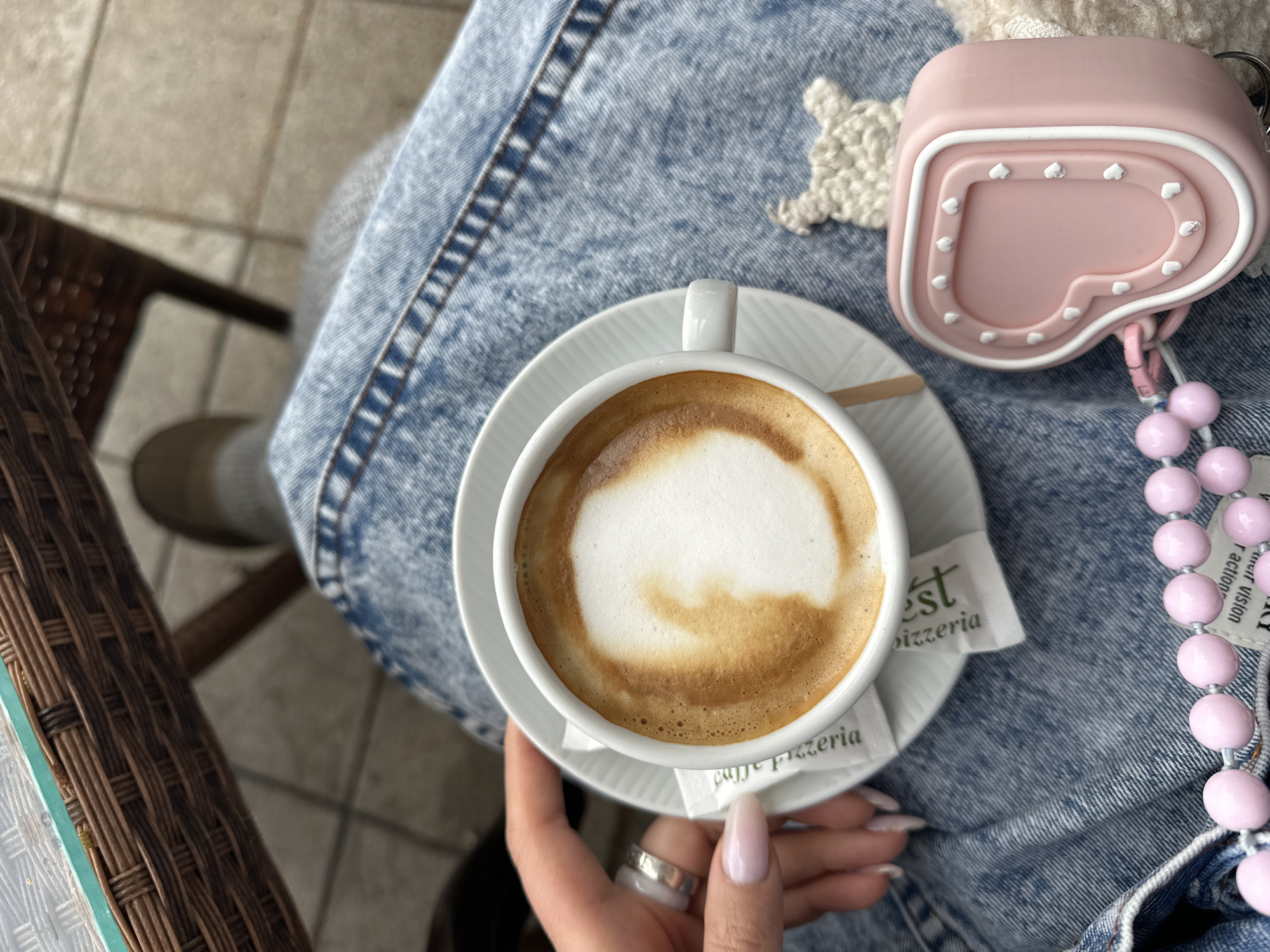 A high-angle, medium shot shows a person sitting at a table holding a cup of coffee. The person is wearing blue jeans. On the jeans, next to the cup, is a pink heart-shaped object with white hearts along the border. A string of pink beads is attached to this object. Two sugar packets with "EST pizzeria" printed on them are near the coffee cup. The hand holding the coffee has long, manicured nails and a silver ring. The background consists of the corner of a wicker chair and tiled flooring. The location is presumed to be a cafe or pizzeria in Bar, Montenegro, based on the sugar packet text.