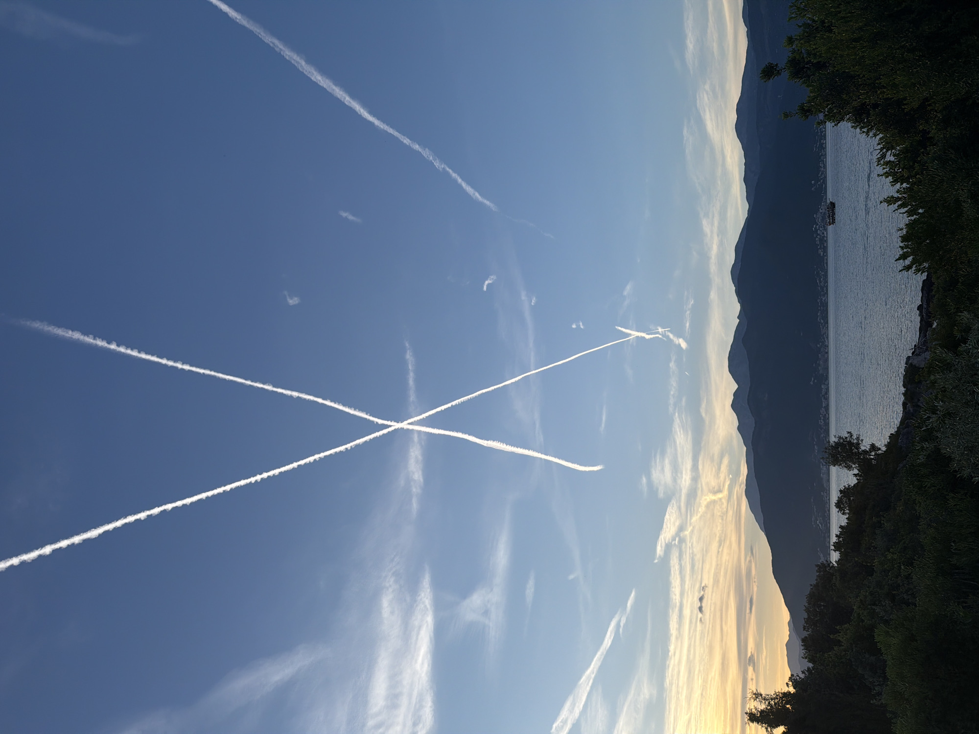 The image shows a landscape with a view of water, possibly a lake or sea, with mountains in the background, likely taken near Gošići, Montenegro. The foreground contains greenery, suggesting a forested or vegetated area. The sky is blue with several prominent contrails from aircraft, some intersecting to form an "X" shape. The sun is setting or rising on the right side, casting a warm glow. A boat can be seen in the water.