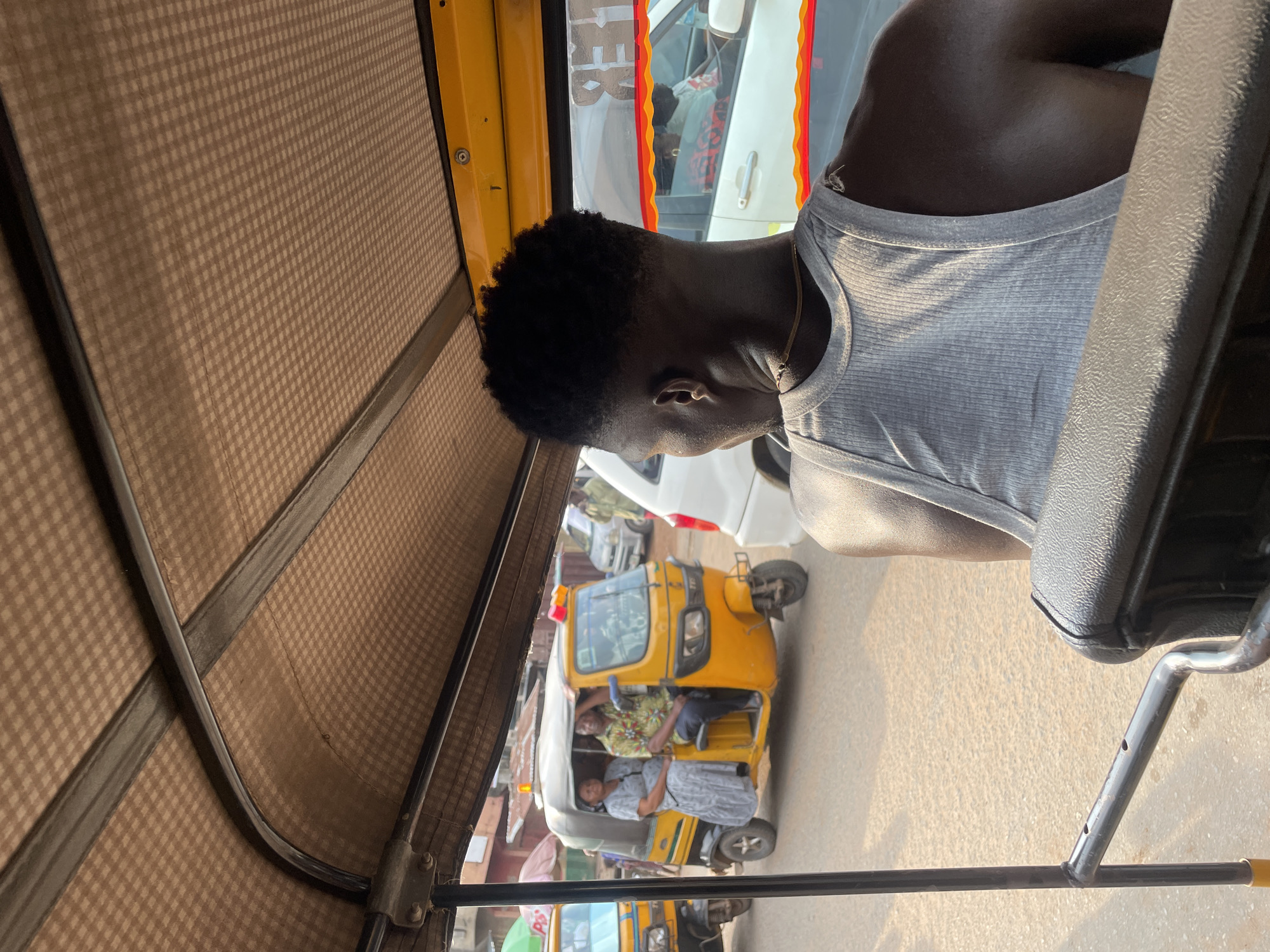 The image presents a forward-facing view from inside a yellow motorized rickshaw (tuk-tuk), likely in an urban street environment in an unknown city. The perspective is from the rear passenger area, looking over the shoulder of the driver.
In the immediate foreground, the back of a male driver is visible. He has dark skin, short, dark, textured hair, and is wearing a light grey ribbed tank top. A thin gold-colored chain is visible around his neck. His right shoulder and upper arm appear muscular and slightly glistening. The black padded backrest of the rickshaw's passenger seat is partially visible in the bottom right, and a black metal support bar with two small circular holes extends horizontally from a vertical post on the left. The roof interior of the rickshaw is visible above, featuring a light brown fabric with a small checkered pattern, supported by dark metal frames and bars.
The scene ahead depicts a street with other vehicles. In the mid-ground, another yellow motorized rickshaw, identified by "TVS" branding on its front, is prominently featured. This rickshaw has a light-colored canvas roof and features small orange and white beacon lights on its top. Inside, a dark-skinned male driver wearing a colorful patterned short-sleeved shirt and dark trousers is seated, his right arm resting on the vehicle's side. A dark-skinned female passenger wearing a light grey or blue patterned long dress is seated to his left.
To the left of the prominent yellow rickshaw, another similar yellow rickshaw is partially visible, mostly obscured by the black support bar of the foreground vehicle. To the right, a white sedan is partially visible and also reflected in what appears to be a window or mirror of the yellow rickshaw ahead. In the distant background, a small grey van or car is visible.
The street surface appears to be a light brown, possibly compacted dirt or unpaved asphalt, illuminated by strong sunlight, creating subtle shadows. On the far left edge, a sliver of a green umbrella or awning and some corrugated metal roofing suggest informal roadside structures or stalls. The overall setting is a bustling street during daylight hours.