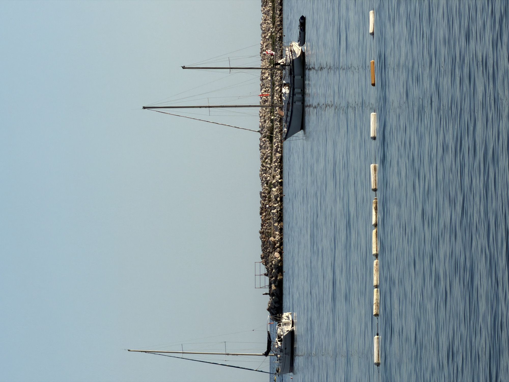 The image presents a serene coastal scene, likely in a marina or harbor, under a clear, light blue sky. Two sailboats are visible on calm, reflective blue water adjacent to a rocky breakwater.
The most prominent sailboat, positioned towards the center-right of the frame, is a monohull with two masts and rigging, suggesting it's a ketch or schooner. It appears to be moored or docked alongside the rocky breakwater. Small red flags or pennants can be seen on its mast, and its clear reflection shimmers on the water's surface. Further to the left, another sailboat, a monohull with a single mast, is partially visible near the far end of the breakwater.
To the right of the breakwater, a straight line of cylindrical objects floats in the water. Most are white, but one in the middle is orange, possibly serving as buoys or fenders. The water throughout the scene is remarkably still, showing subtle, elongated reflections and ripples. The rocky breakwater itself forms a natural boundary, separating the immediate mooring area from a wider expanse of water where the buoys are situated. The sky is a uniform, pale blue, indicative of bright, clear weather. No people or visible text are discernible. The overall impression is one of peaceful tranquility in a daylight setting. The location context indicates this scene is in Budva, Montenegro.