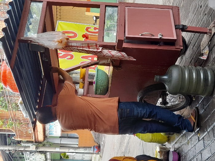 A man in an orange shirt and jeans stands next to a food cart on a brick sidewalk. He appears to be adjusting or working on the cart's structure. The cart has a corrugated metal roof, a glass display case, and a closed compartment with a handle. A large plastic bottle and a bicycle wheel are visible beneath the cart. A colorful banner and various containers are visible within and around the cart. Buildings and greenery are in the background.