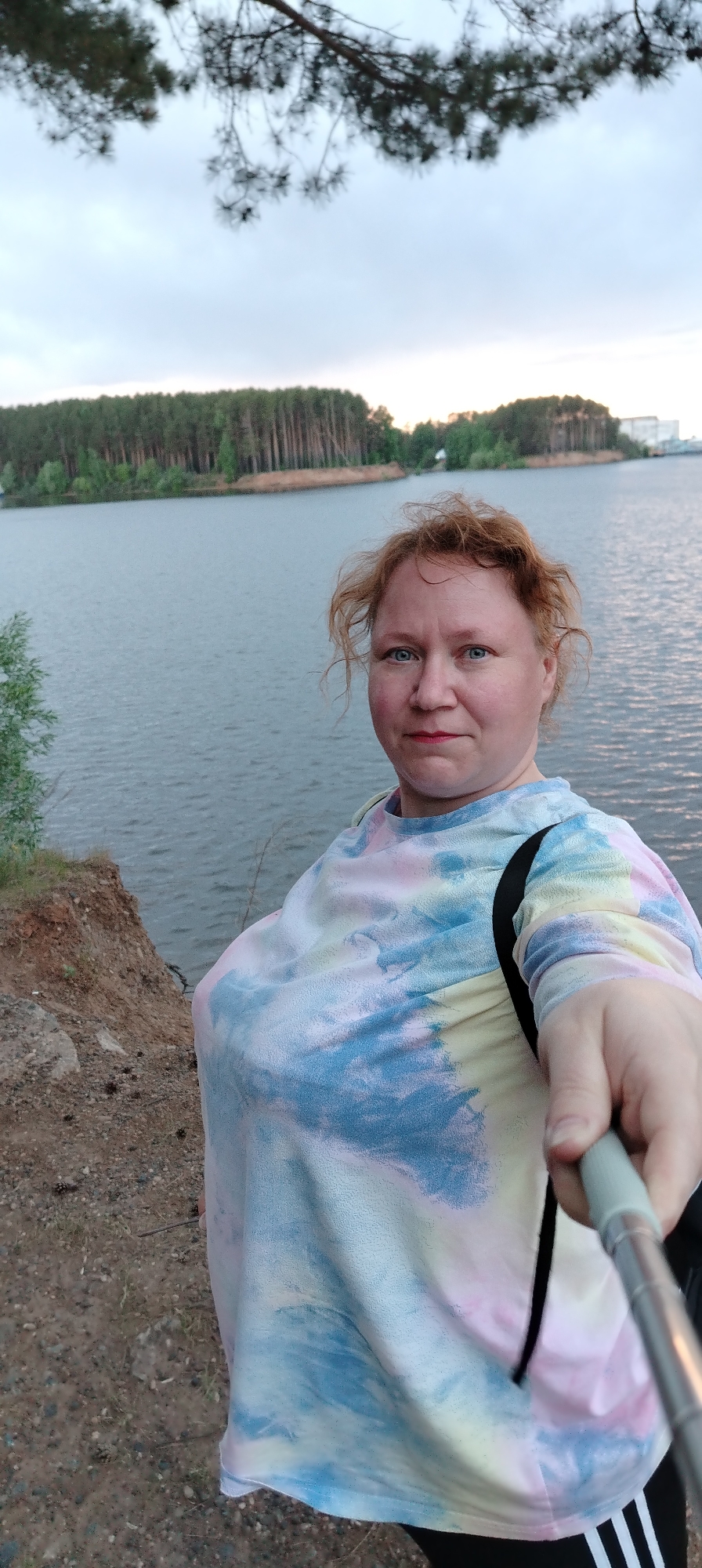 A woman is taking a selfie with a selfie stick. She is standing on a cliff overlooking a body of water with a forested area and a building in the background. The location appears to be in the city of Chaikovsky, Russia. The woman is wearing a tie-dye shirt and Adidas track pants. There is foliage above, a body of water and a forest in the background.