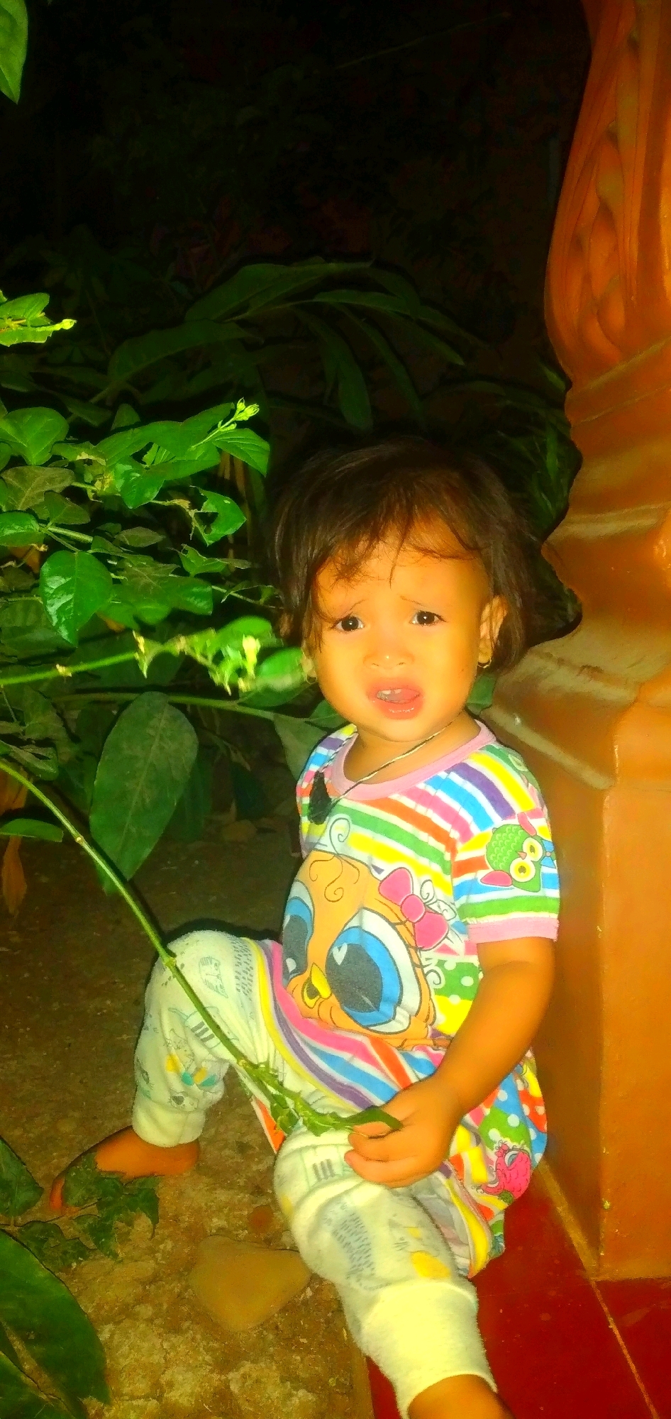 A young child, appearing to be of Southeast Asian descent, is sitting on the ground amidst foliage and an orange-colored structure, possibly a wall or pillar. The child is wearing a brightly colored, striped shirt with a cartoon character graphic on the front and light-colored pants with a pattern. The child's hair is dark and appears to be styled short. They are holding a green stem in their right hand and have a somewhat distressed or surprised expression on their face, with their mouth slightly open. The setting appears to be outdoors, possibly in a garden or courtyard, given the presence of plants and the ground material. The lighting suggests it might be dusk or night, with artificial illumination casting a warm glow on the scene. The orange structure on the right side of the image provides a stark contrast to the greenery and the child's attire. The overall impression is of a candid, possibly impromptu photograph capturing a moment with the child in an outdoor environment in Rahong, Indonesia.
