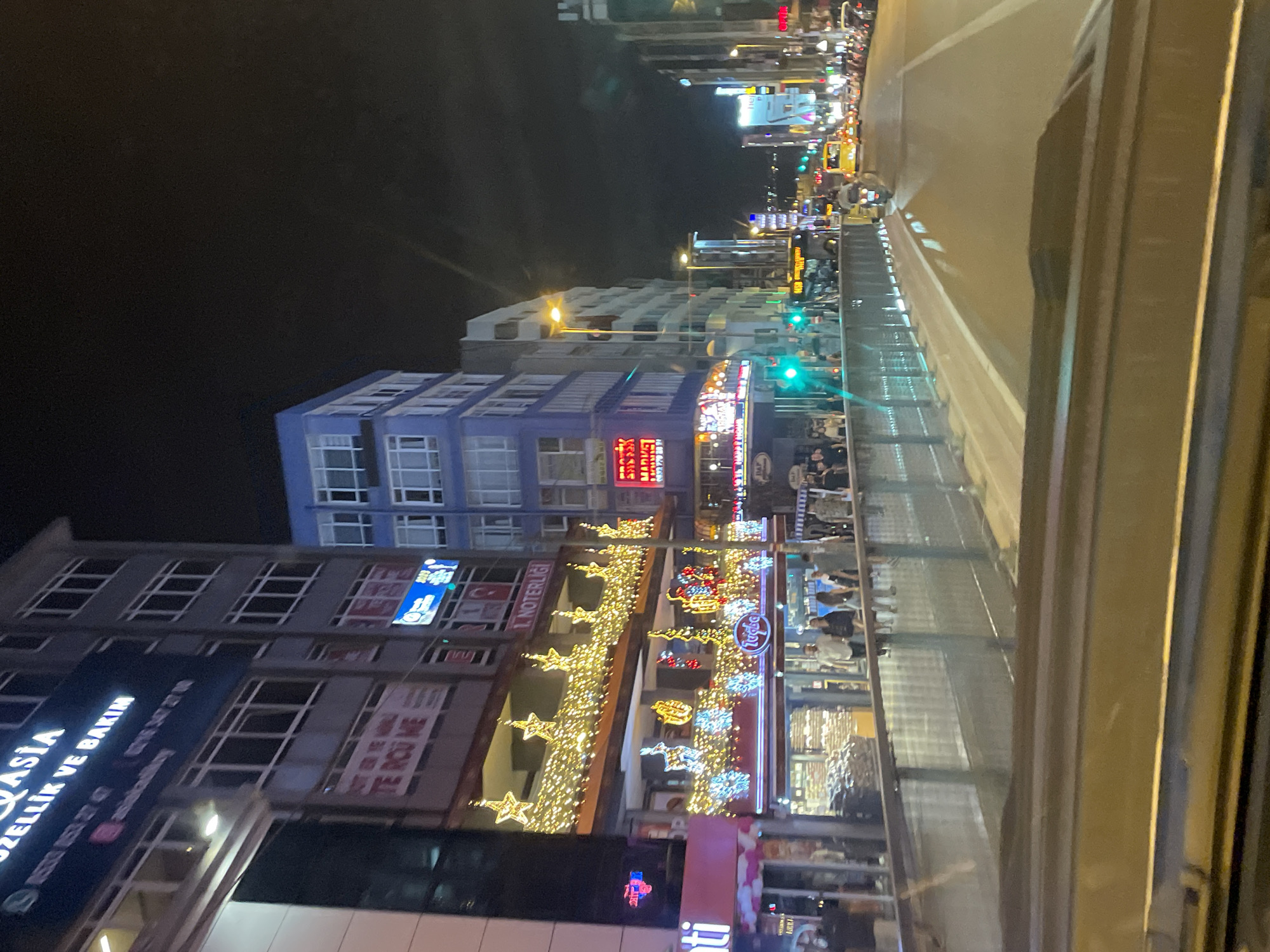 The image depicts a night scene in Kadıköy, Turkey, specifically along a street lined with buildings and illuminated by streetlights and commercial signage. In the foreground, a portion of a vehicle's interior, likely a dashboard or hood, is visible. A pedestrian walkway with a railing separates the street from the buildings.
On the left, a multi-story building displays various signs, including one that reads "ASIA" in large letters, followed by "ÖZELLIK VE BAKIM". Below this, there is a sign with a pink background that appears to be for a business. Further down the street, a building is adorned with bright, festive lights, including stars, reindeer, and snowflakes, suggesting a holiday or celebratory decoration. Businesses are visible at street level, with people walking on the sidewalk.
To the right, other buildings are illuminated, with prominent commercial signs. Traffic is present on the street, with yellow taxis and other vehicles visible. A motorcycle is seen moving along the road. The overall atmosphere is urban, with a sense of activity and commercial vibrancy despite the late hour. The sky is dark, indicating nighttime.