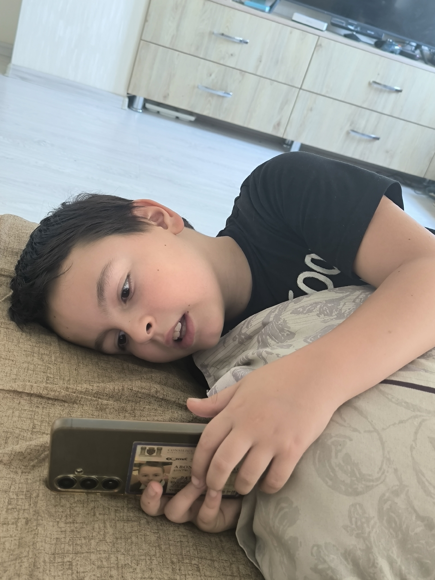 A young boy, appearing to be of East European descent, lies on a tan-colored sofa, propped up by a pillow. He is wearing a black t-shirt with white lettering. The boy is holding a smartphone in his right hand, with a small identification card visible on the back of the phone. The card features a photograph of the boy, along with text that includes "CONSILIUL" and "ABON".
The boy's head is turned to his left, and he is looking towards the phone. His mouth is slightly open, as if he is speaking or reacting to something on the screen. His left arm is extended, supporting his head on the pillow.
In the background, a light-colored wooden dresser with silver handles is visible. The floor is light-colored laminate. The lighting in the room is bright, suggesting it is daytime and natural light is entering from an unseen source.
The scene appears to be an indoor residential setting within the city of Chișinău, Moldova, as indicated by the script on the identification card.