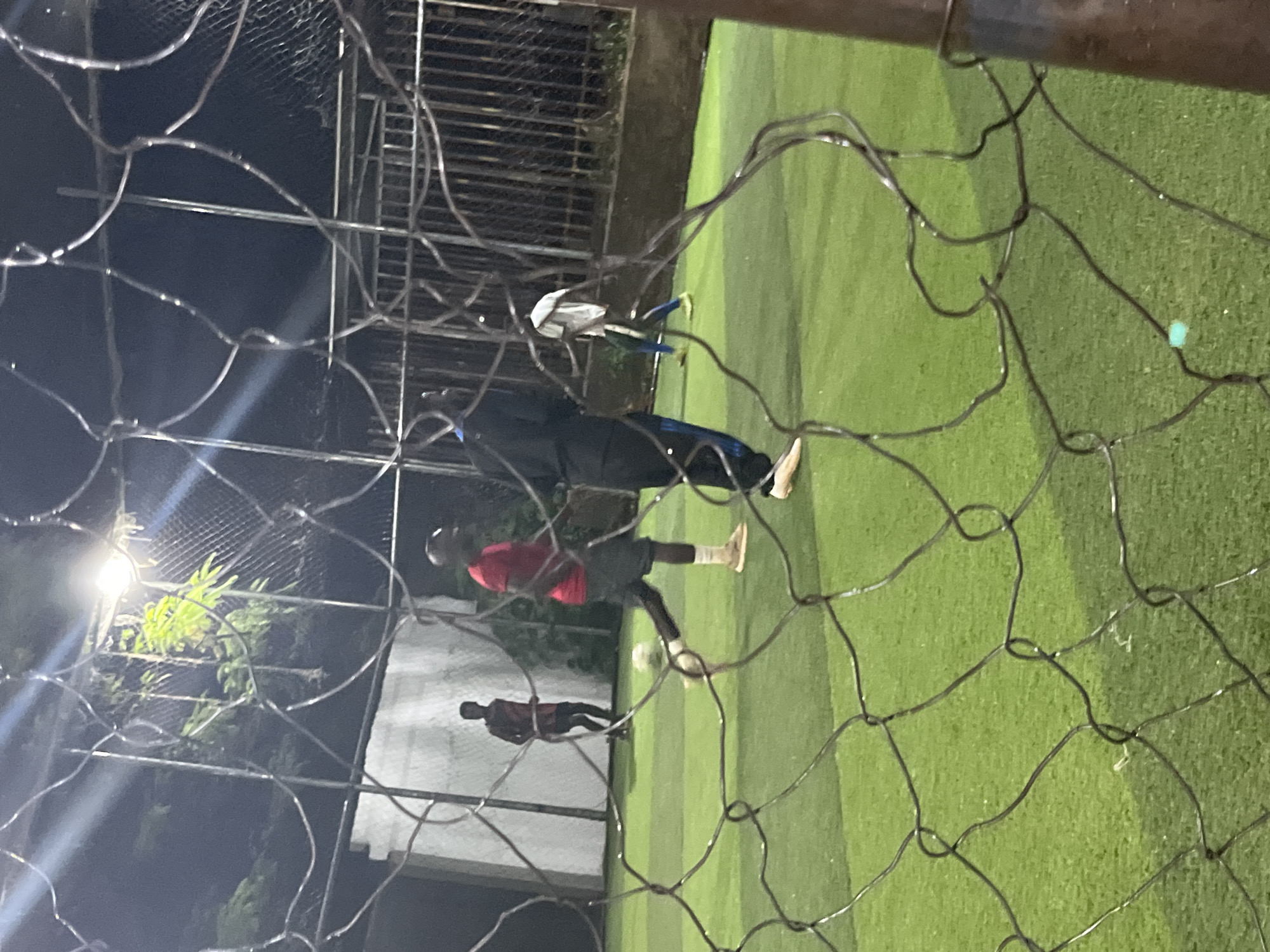 The image depicts a group of individuals playing soccer on an artificial turf field at night, possibly at the Old GRA soccer pitch in Jos, Nigeria. The scene is illuminated by overhead lights, casting a slightly dim and grainy effect on the photograph. The foreground is partially obscured by a dark mesh fence, typical of a sports enclosure.
In the mid-ground, several men are engaged in the game. One man in a red t-shirt and dark shorts is prominently positioned, appearing to be mid-stride with a soccer ball near his feet. Another individual in a black tracksuit with blue stripes is also visible. Further back, a person in a white t-shirt and blue shorts is running, and another in a dark t-shirt and shorts is standing. The background shows a white wall on the left and a metal fence behind the field. Trees are visible beyond the fence, and the overall impression is of an active sports facility after dark.