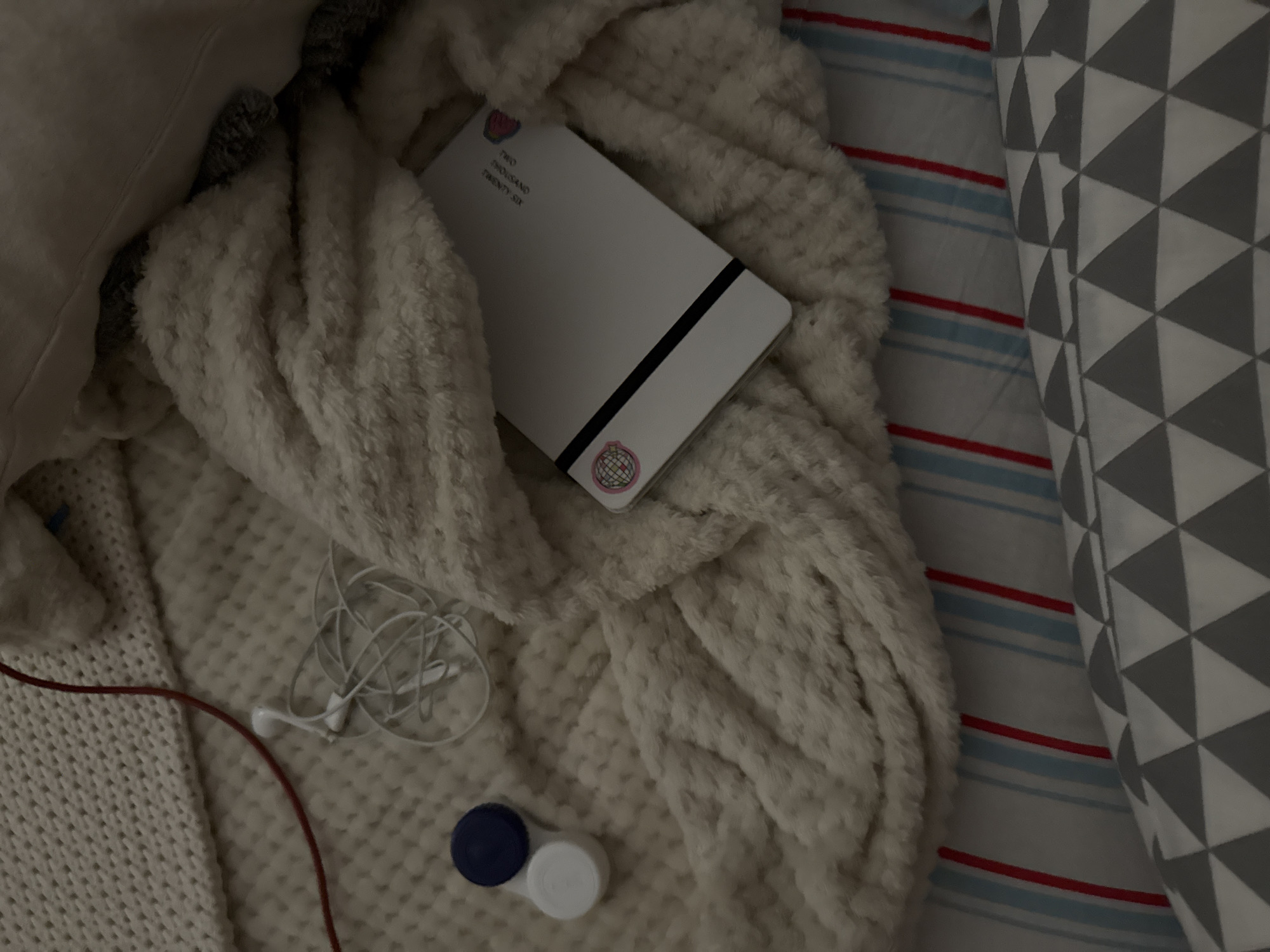 The image shows a close-up, overhead view of a white fuzzy blanket on a bed. A white notebook with a black stripe and two circular logos is tucked into the blanket. To the left of the notebook, on a textured white surface that appears to be a different blanket or throw, are a pair of white earbuds and a white contact lens case with a blue lid. The bed linens below the fuzzy blanket have a pattern of thin blue and red stripes. The overall scene is indoors and dimly lit. There is no information to suggest the location is in the city of Izobilnyy, Russia.