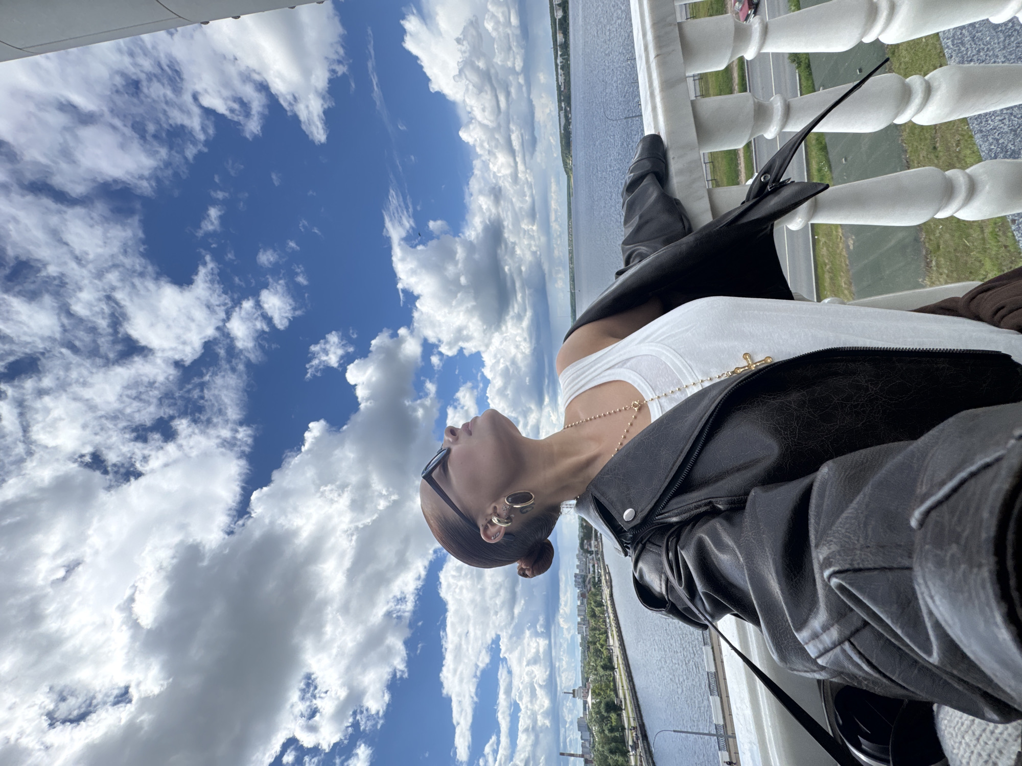 A woman stands on a balcony with white balusters, overlooking a body of water with a city in the background, likely Izhevsk, Russia, based on available visual information. She wears a white tank top, a black leather jacket, and trousers. She has multiple earrings and a necklace with a cross pendant. The sky is blue with scattered clouds.