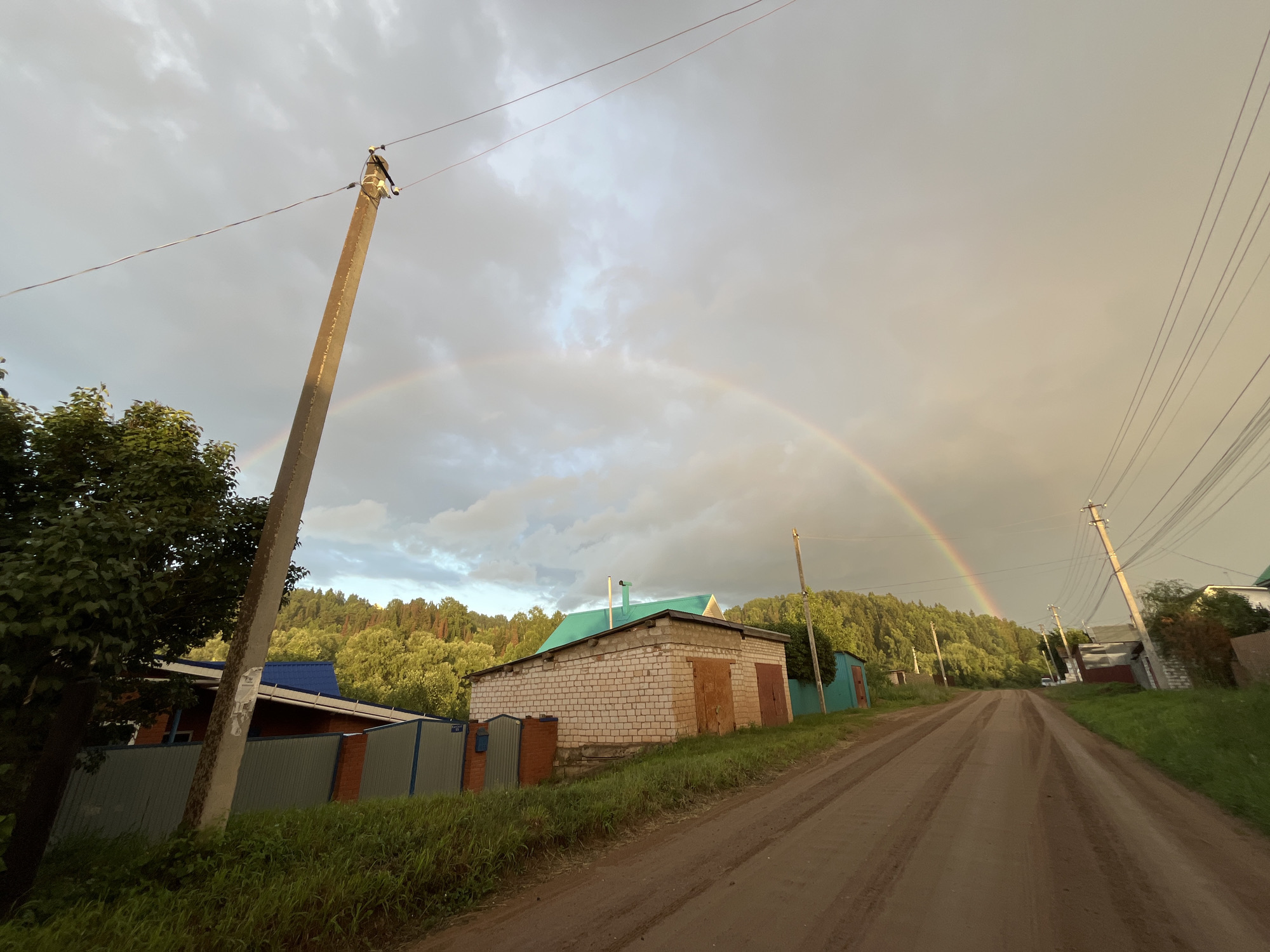 The image depicts a rural scene in Olkhovka, Russia, under a cloudy sky with a rainbow arching across it. A dirt road runs through the center of the frame, leading towards a wooded hillside. On either side of the road are several buildings, including a brick structure with two garage doors, and other smaller outbuildings. Power lines and utility poles are visible throughout the scene. The vegetation is green and lush, suggesting a temperate climate. The time of day appears to be late afternoon or early evening, given the quality of light. The overall mood is peaceful and serene, with the rainbow adding a touch of wonder to the landscape. There is no visible text in the image.