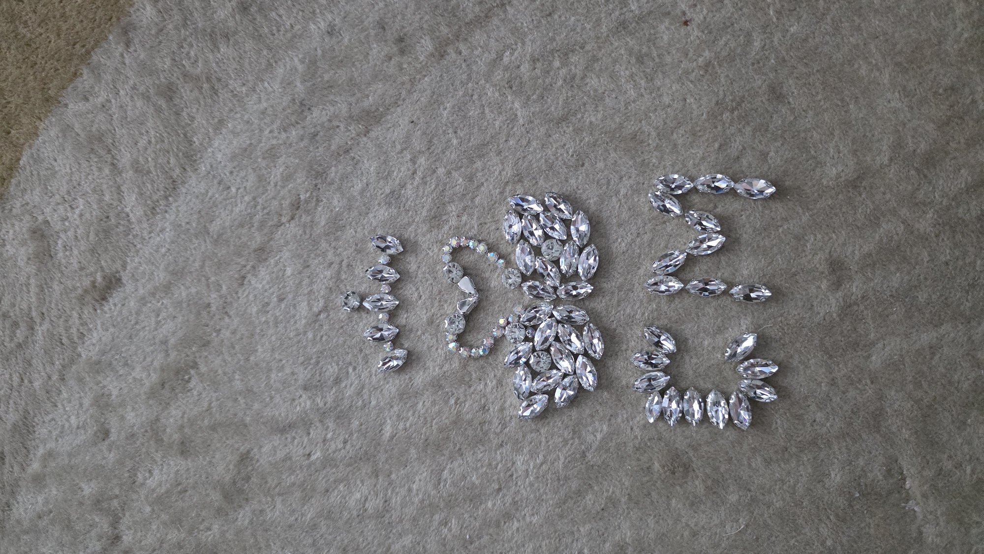 The image presents an artistic arrangement of clear, faceted, marquise-cut rhinestones and some iridescent round beads, meticulously placed on a light beige, textured carpet or rug. No people are visible in the scene, which appears to be set indoors.
The rhinestones are arranged to form a sequence of characters. From left to right, the first character is the numeral "1", formed by a vertical stack of marquise rhinestones. Following this, a more elaborate design features a central cluster of marquise rhinestones from which two curved lines of iridescent beads extend, resembling a stylized "S", a butterfly, or a decorative numeral "0". Next in the sequence is the numeral "2", also formed from marquise rhinestones. The final character, positioned below and to the right of the "2", is the numeral "3", created by a curved arrangement of marquise rhinestones. Interpreting the stylized shape as a "0", the full sequence of visible text spells out "1023".
The overall activity depicted is the creative arrangement of decorative elements. The rhinestones, with their reflective surfaces, catch the light, adding a sparkling quality to the composition. The setting offers no specific cues regarding time of day or weather, as it is an indoor shot on a soft, fluffy surface. No emotions are conveyed, as the image focuses solely on the arranged objects. The location context for the image is Blagoevgrad, Bulgaria, but this is not visually evident in the media file itself.