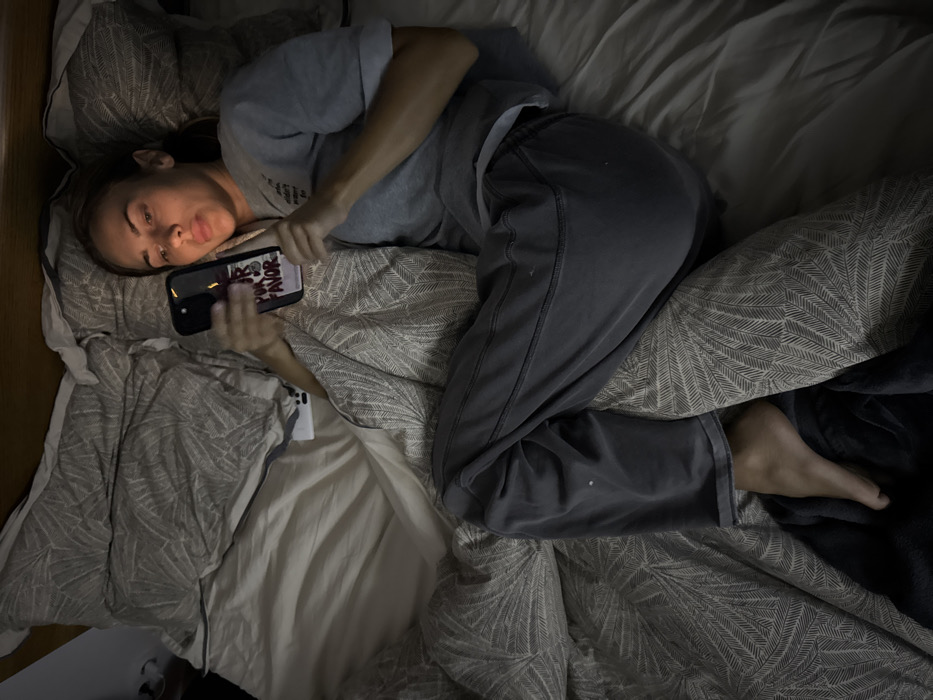 A person is depicted lying on their left side in a bed, actively engaged with a smartphone. They are dressed in a light grey short-sleeved t-shirt and dark grey sweatpants. The bed is adorned with patterned bedding, featuring a repeated leaf or fern-like motif in varying shades of grey, with a lighter, plain sheet visible underneath. The lighting in the room is dim, suggesting it is either nighttime or a poorly lit environment.
The person's head rests on a pillow, and their face is turned towards the viewer, displaying a neutral expression with slightly pursed lips. They are holding a black-cased smartphone with both hands, positioned directly in front of their face. Visible text on the phone screen clearly reads "POR FAVOR," with additional, partially obscured text above it that appears to be "SI" or "SO," set against a background featuring red and white elements. A white cable or cord is visible near the person's right hand, lying on the bed. The individual's bare feet are partially visible towards the bottom right of the frame, tucked under a dark blanket. This intimate scene takes place in Warsaw, Poland.