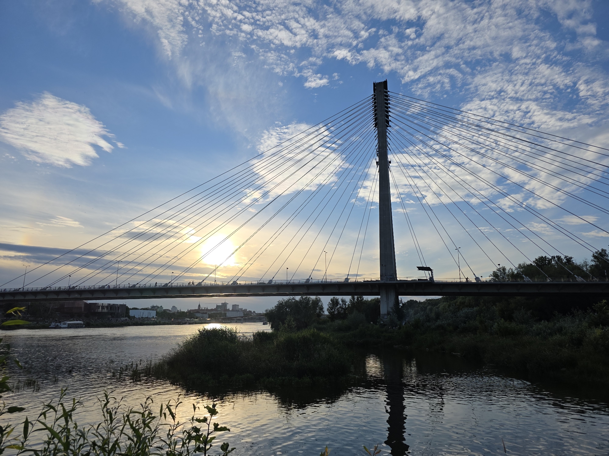 This is a wide shot of the Swietokrzyski Bridge over the Vistula River in Warsaw, Poland. The sun is setting, casting a warm glow on the sky and water. The bridge, a suspension bridge with a single pylon, is in the foreground, with its cables stretching across the sky. The river is calm and reflects the light. Trees and vegetation line the banks of the river. A few buildings and a boat are visible in the distance. There are no discernible people in the frame, but there are silhouettes of what appear to be individuals on the bridge walkway.