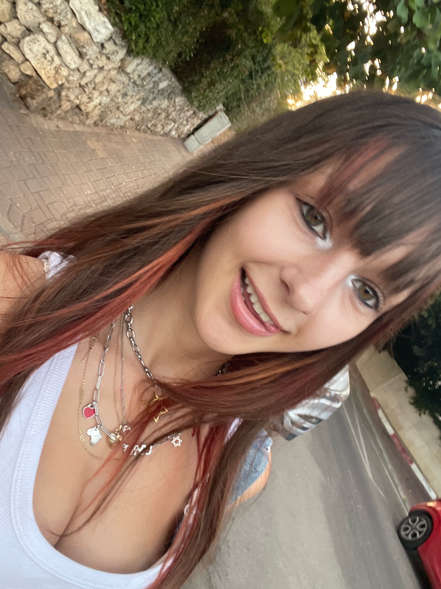 A young woman is taking a selfie on a street in Raanana, Israel. She is wearing a white tank top and multiple layered necklaces. Her hair is long and brown with reddish highlights. She has winged eyeliner and is smiling with her tongue slightly out. The background shows a stone wall, a paved sidewalk, and some greenery. A red car is visible in the lower right corner of the image.