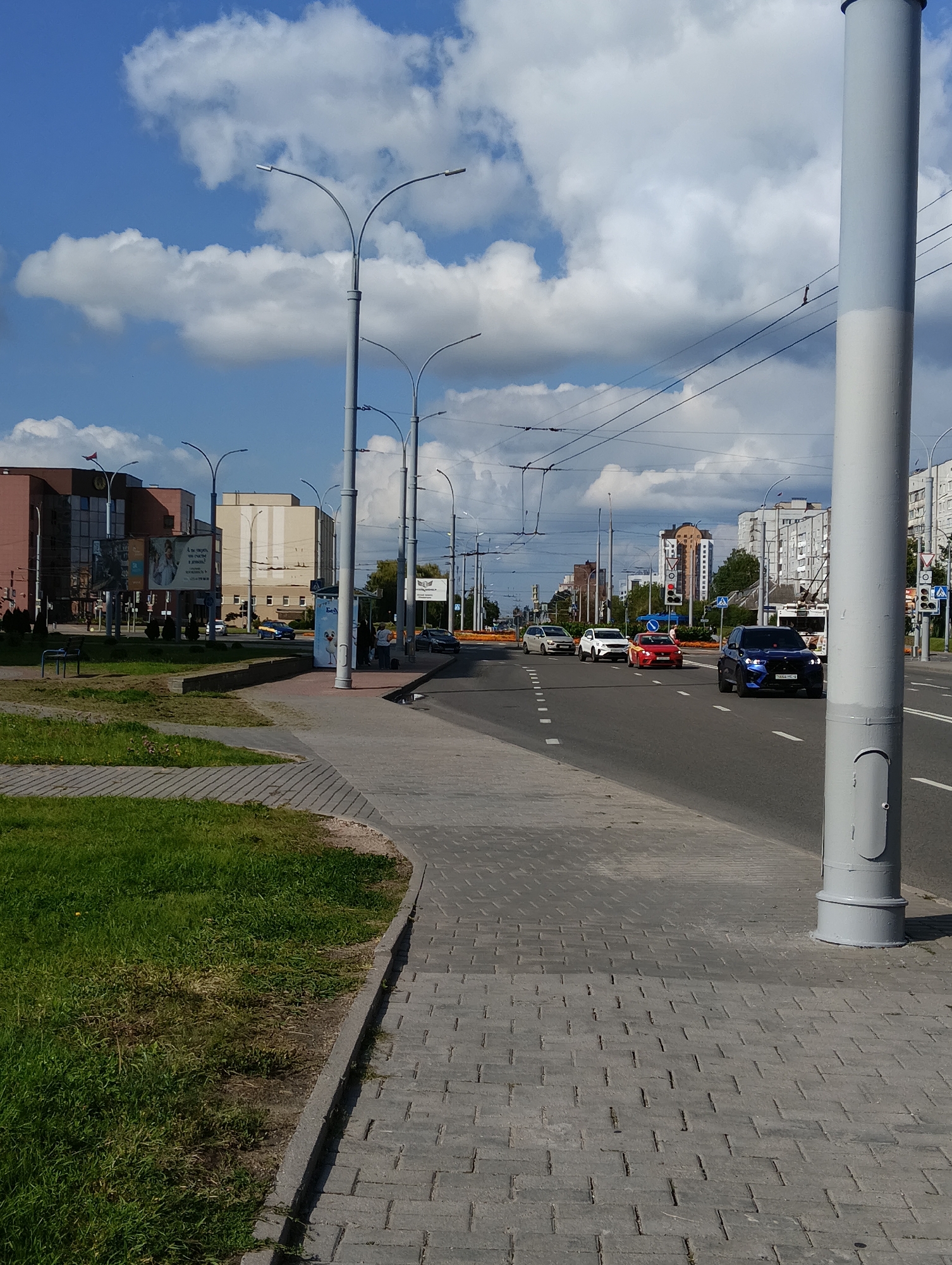 The image depicts a city street scene in Hrodna, Belarus, on a sunny day with scattered clouds. In the foreground, a paved sidewalk with a pattern of rectangular bricks runs along the left side of the image. Adjacent to the sidewalk is a strip of green grass. A large, light gray cylindrical pole, possibly a lamppost or utility pole, is prominently positioned in the right foreground.
The street itself has multiple lanes of asphalt road with white lane markings. Several vehicles are visible: a dark blue SUV in the right lane, a red sports car to its left, a white sedan, and a lighter colored van further down the road. Traffic lights and street signs are present, along with overhead power lines for trolleybuses.
On the left side of the street, there are buildings, including a multi-story brick building with large windows and what appears to be signage. A bus stop shelter with people waiting is also visible on the sidewalk near the right side of the image. The general environment suggests an urban setting with active traffic and pedestrian activity.