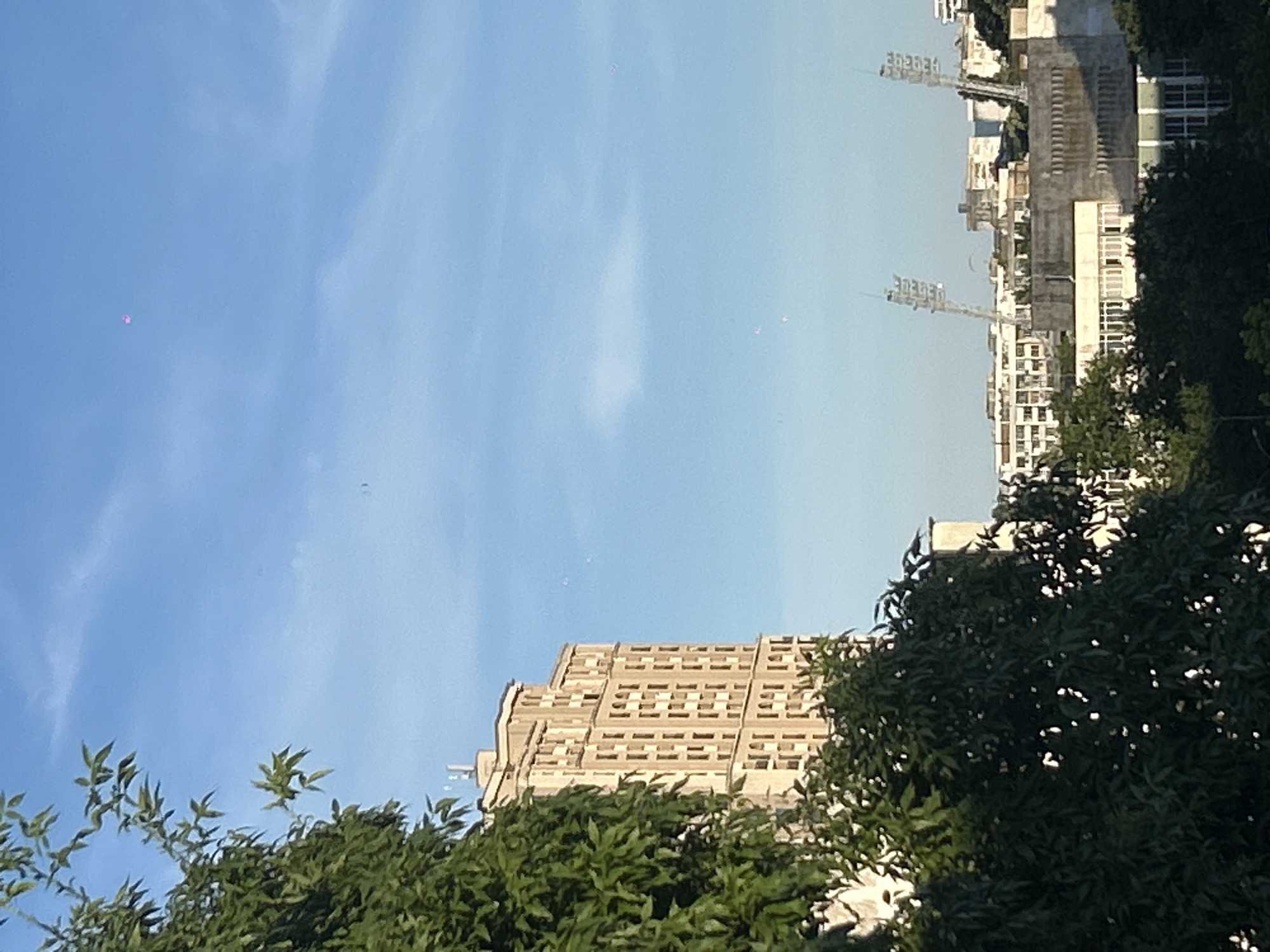 A bright, sunny day in Burgas, Bulgaria. In the foreground, the lush green leaves of a tree obscure much of the left side of the image. Behind the tree, a tall, light-colored apartment building dominates the mid-ground, its facade adorned with numerous windows. In the distance, various other buildings, including what appear to be stadium lights, are visible against a clear blue sky with wispy clouds. A small, pink object, possibly a balloon or drone, is visible high in the sky. There are no people or animals clearly identifiable in the image.