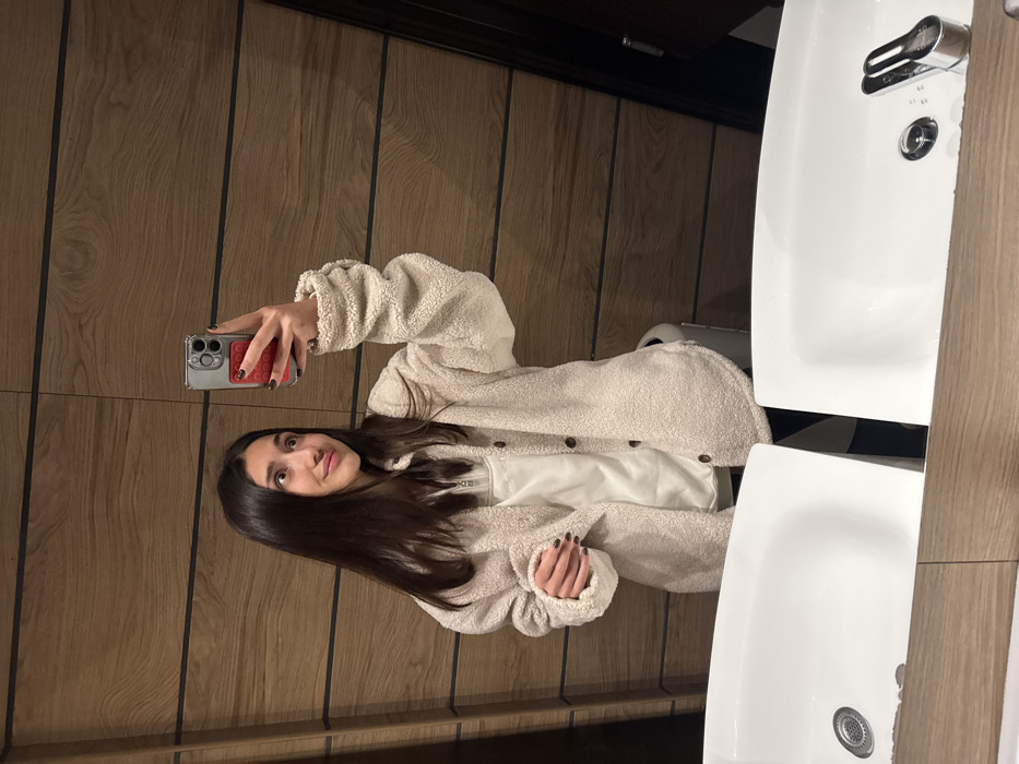 A young woman with long dark hair holds a cell phone, presumably taking a selfie, in what appears to be a public restroom. She is wearing a cream-colored, button-down, fleece-like jacket over a white shirt. The background includes wood-paneled walls and two white sinks with chrome fixtures. The sinks are positioned side by side, suggesting a communal or public restroom space.